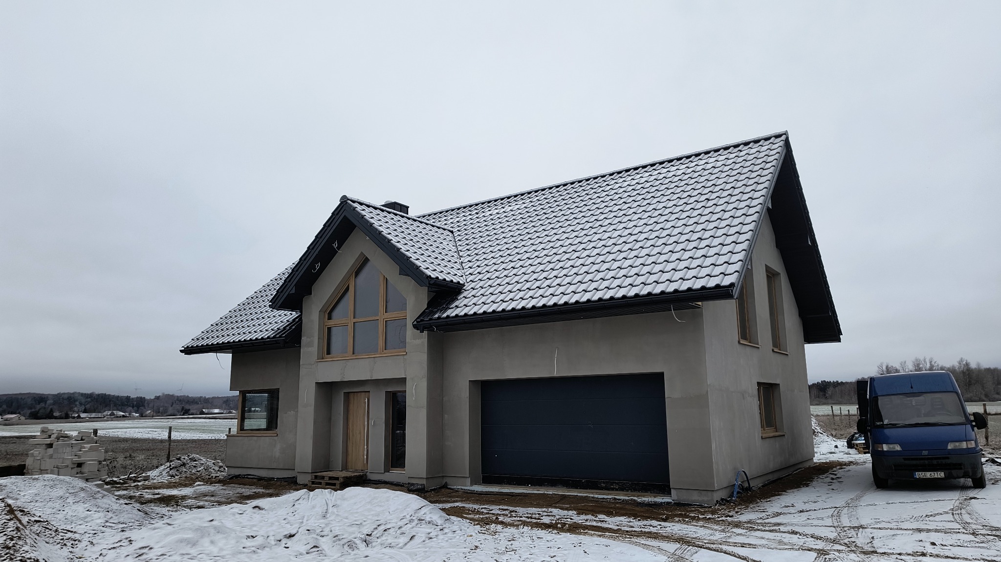 Surowy dom jednorodzinny z szarą elewacją i ciemną dachówką pokrytą śniegiem, widoczny w zimowej scenerii z zaparkowanym vanem w tle.