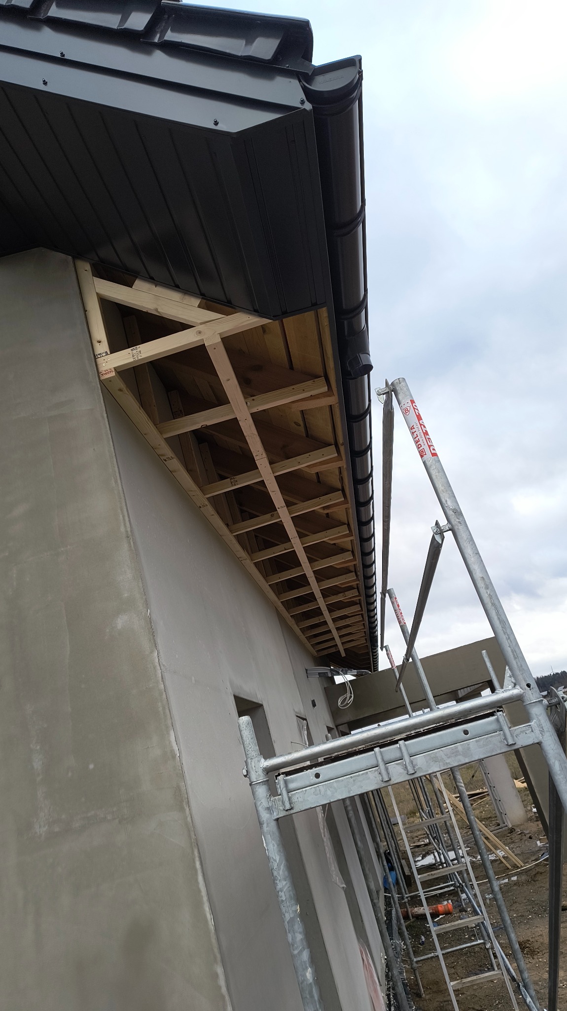 Surowa ściana budynku z nałożoną szarą warstwą, widoczne drewniane elementy konstrukcyjne pod dachem oraz rusztowanie.