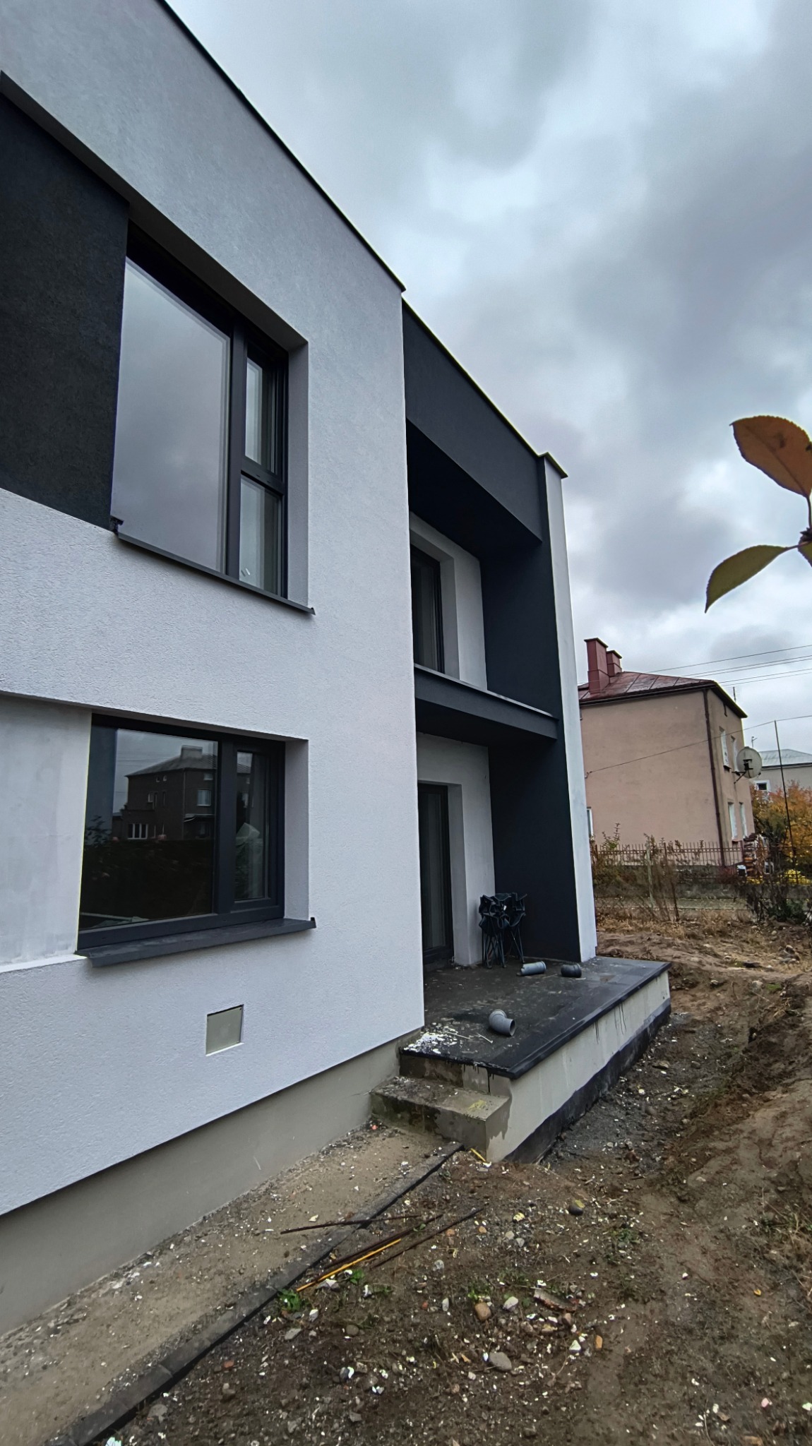 Dwukondygnacyjny dom z białą elewacją i czarnymi akcentami architektonicznymi, widok z boku na niezagospodarowany teren działki i sąsiedni budynek w tle.