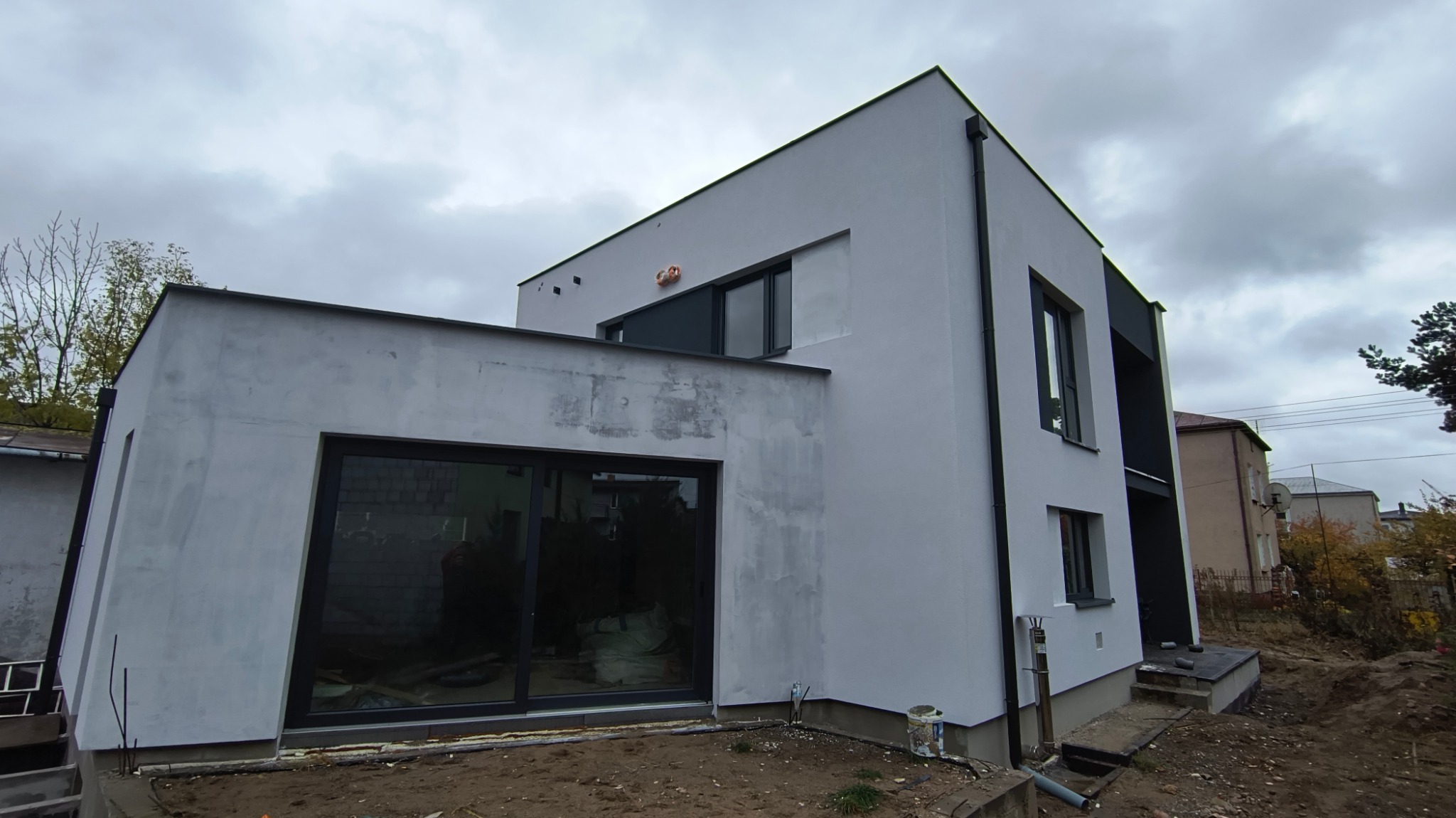 Nowoczesny dom z elewacją w trakcie wykończenia, widoczne ślady po gładzi na części ściany, duże okna z czarnymi ramami, teren wokół domu częściowo przygotowany pod trawnik.