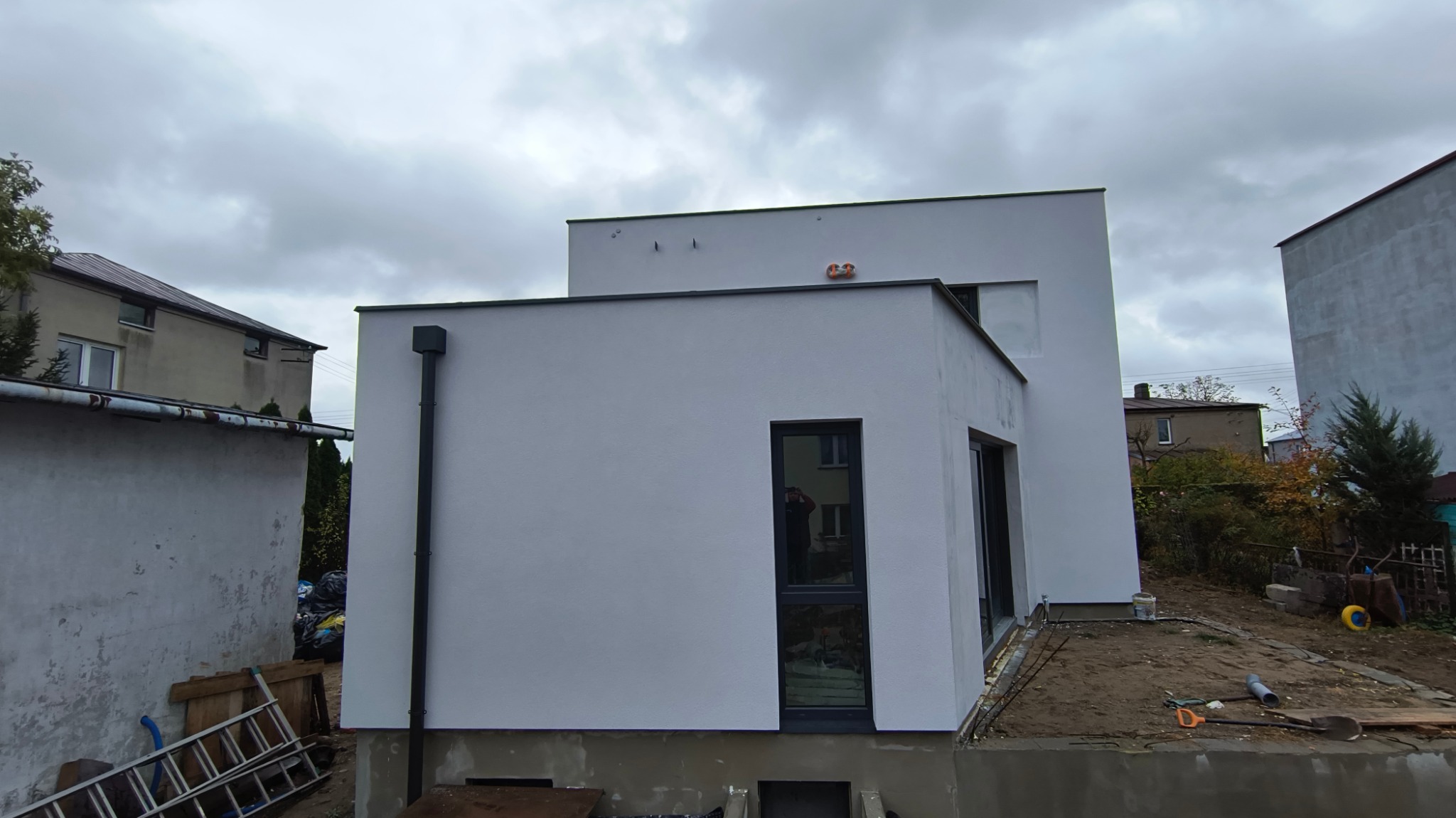 Nowoczesny dom z białą elewacją, widoczny z zewnątrz, z czarnymi ramami okiennymi i rynną, na tle pochmurnego nieba.