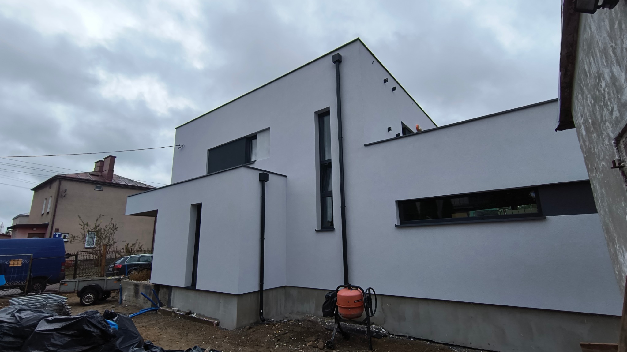 Nowoczesny, biały dom jednorodzinny z płaskim dachem, z czarnymi oknami i rynnami, widziany z boku, z pomarańczową betoniarką na pierwszym planie i sąsiednim budynkiem w tle.