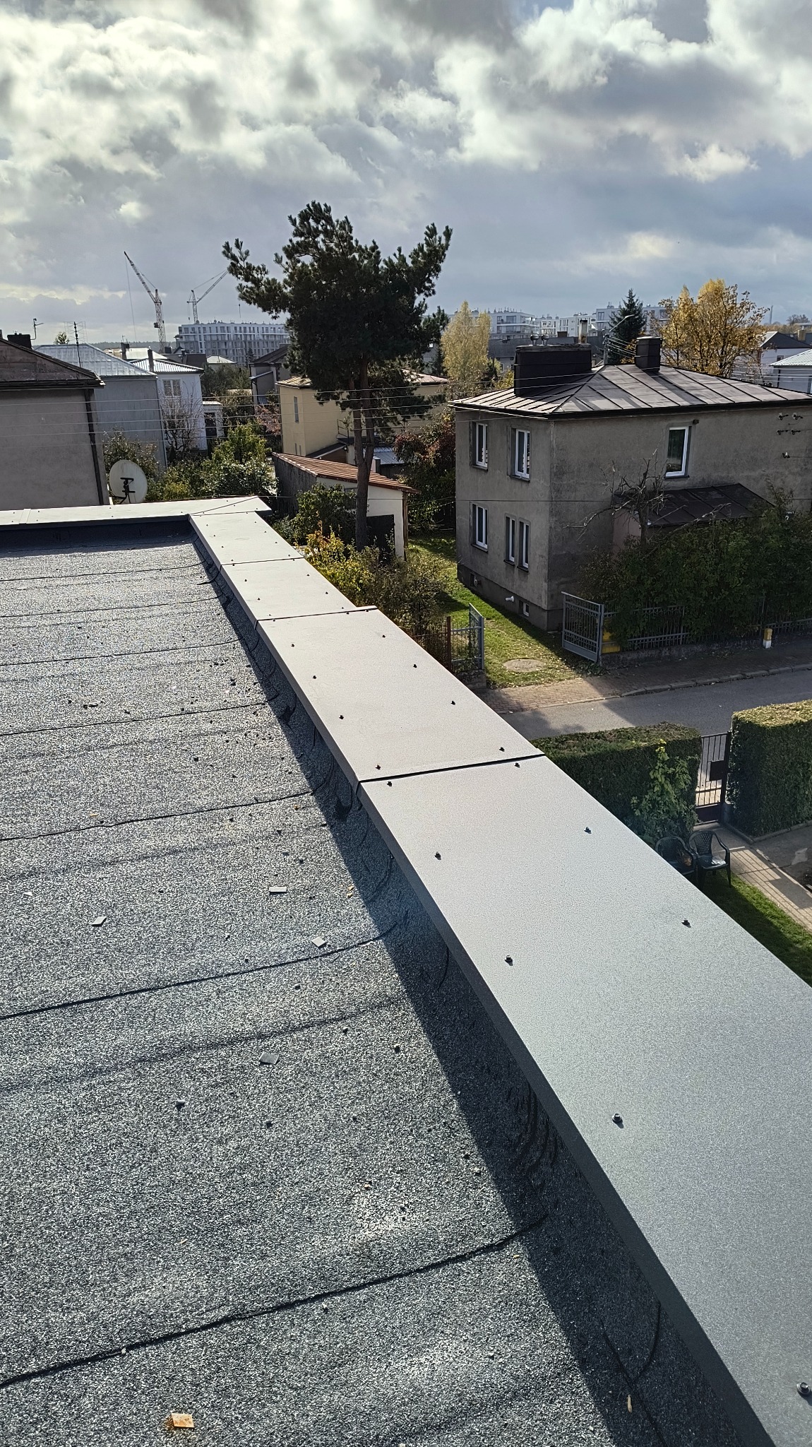 Widok z góry na fragment dachu pokrytego papą z zamontowaną szarą obróbką blacharską, w tle zabudowa miejska i pochmurne niebo.