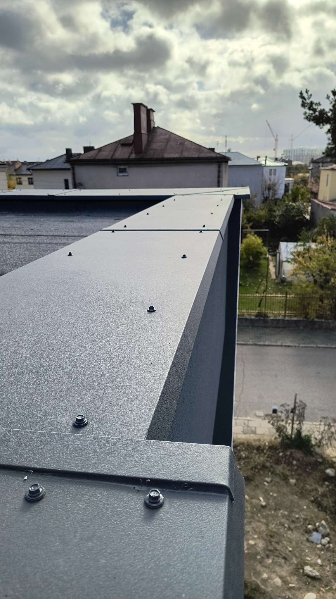 Szczegółowe ujęcie szarej, metalowej obudowy balkonu z widocznymi śrubami mocującymi, na tle zabudowy miejskiej i pochmurnego nieba.