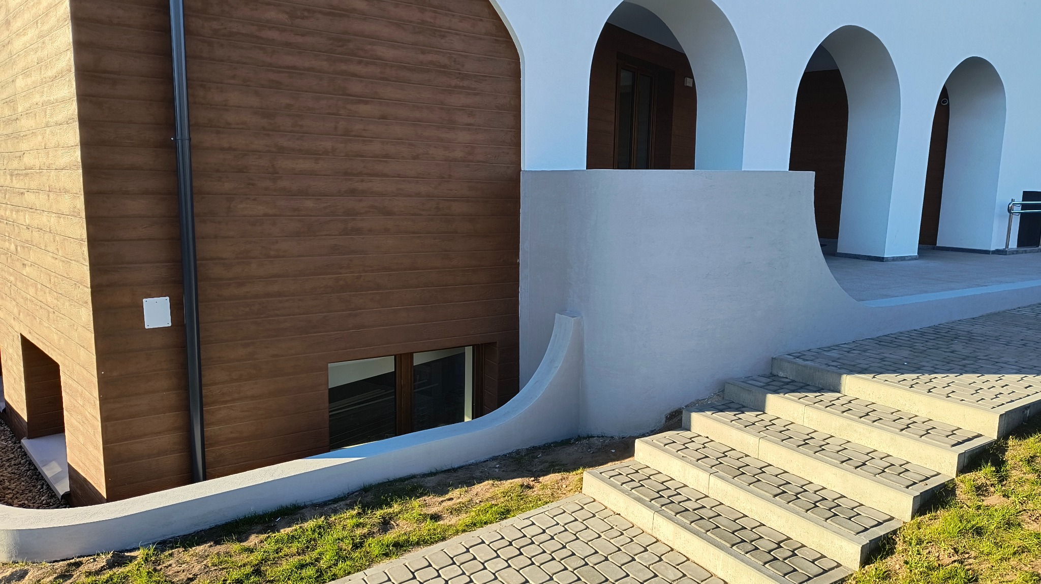 Elewacja budynku z drewnopodobną okładziną, białymi łukami i schodami z kostki brukowej, widok z dołu na jasnym tle.