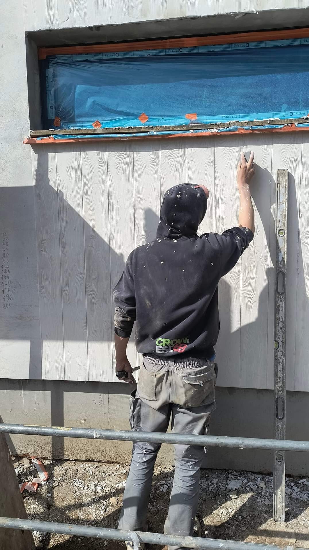 Pracownik w bluzie z kapturem, widziany od tyłu, wygładza szpachlą ścianę budynku z oknem zabezpieczonym folią, obok stoi poziomica.