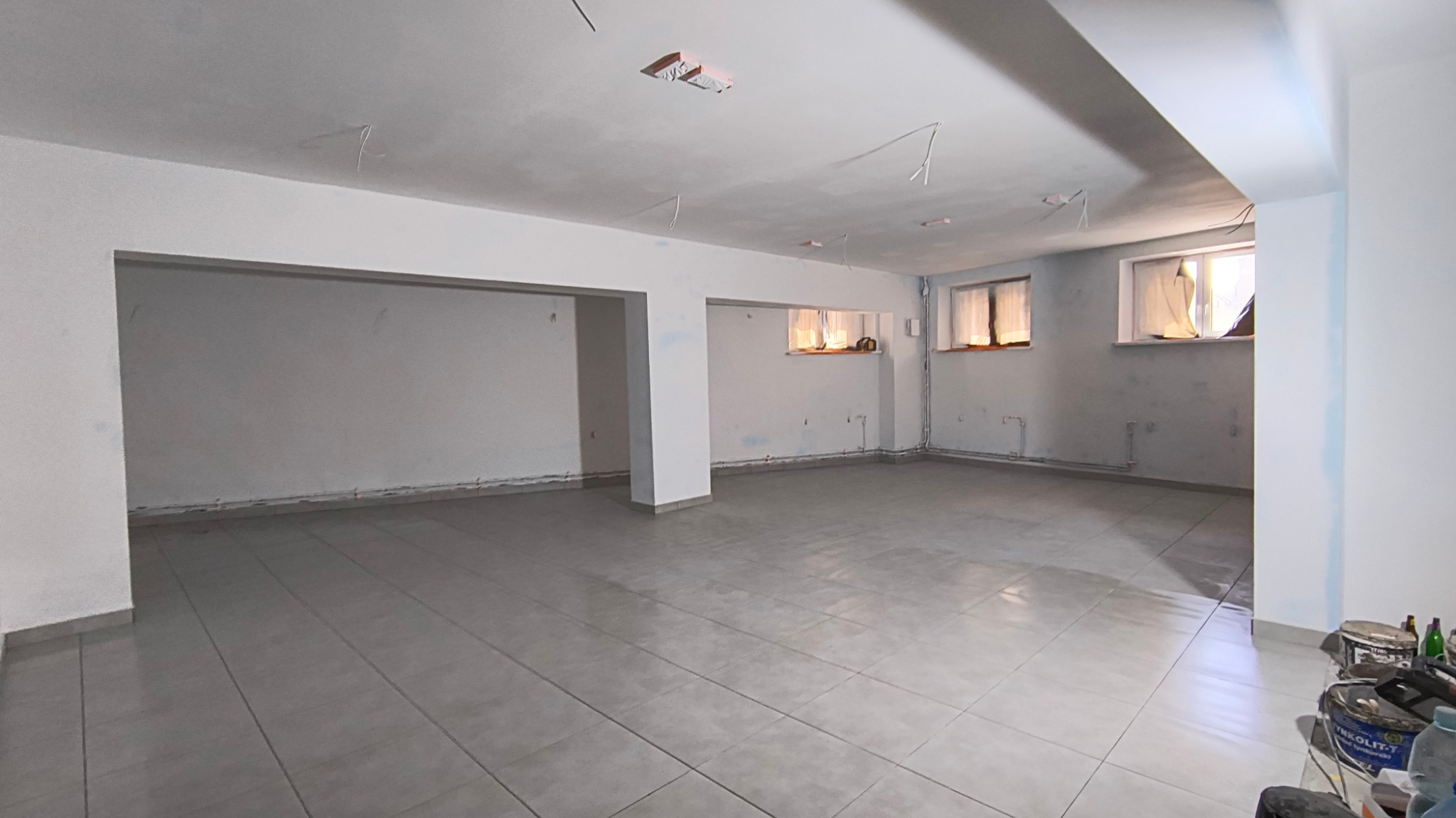 Przestronne pomieszczenie w trakcie remontu, z szarą podłogą z płytek, surowymi ścianami częściowo pomalowanymi na biało, widoczne instalacje elektryczne i rury, oraz pojemniki z farbą w rogu.