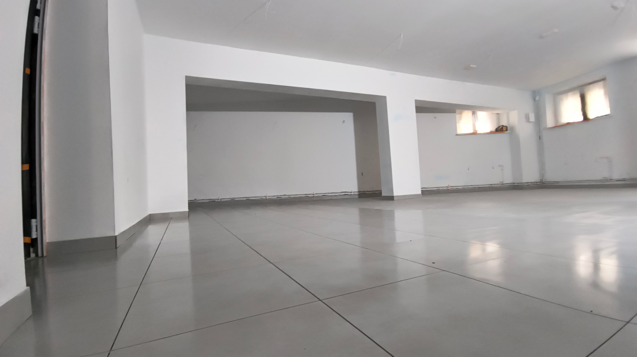 Wnętrze pustego, odnowionego biura z szarą podłogą z płytek, białymi ścianami i widocznymi w oknach promieniami słonecznymi. Widoczne ślady po instalacjach na ścianach.