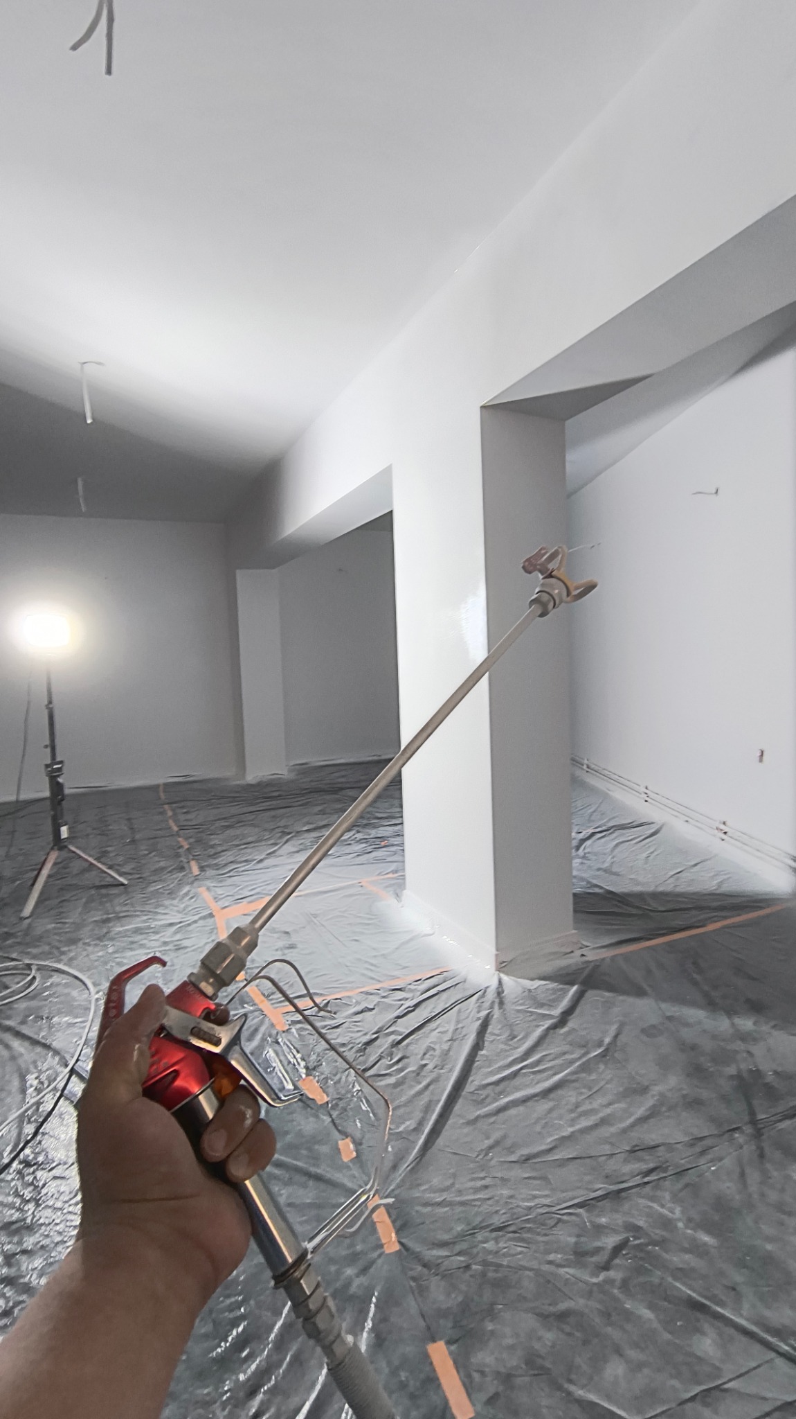 Malowanie ścian w pomieszczeniu biurowym za pomocą pistoletu natryskowego, widoczne zabezpieczenia podłogi folią i oświetlenie halogenowe.