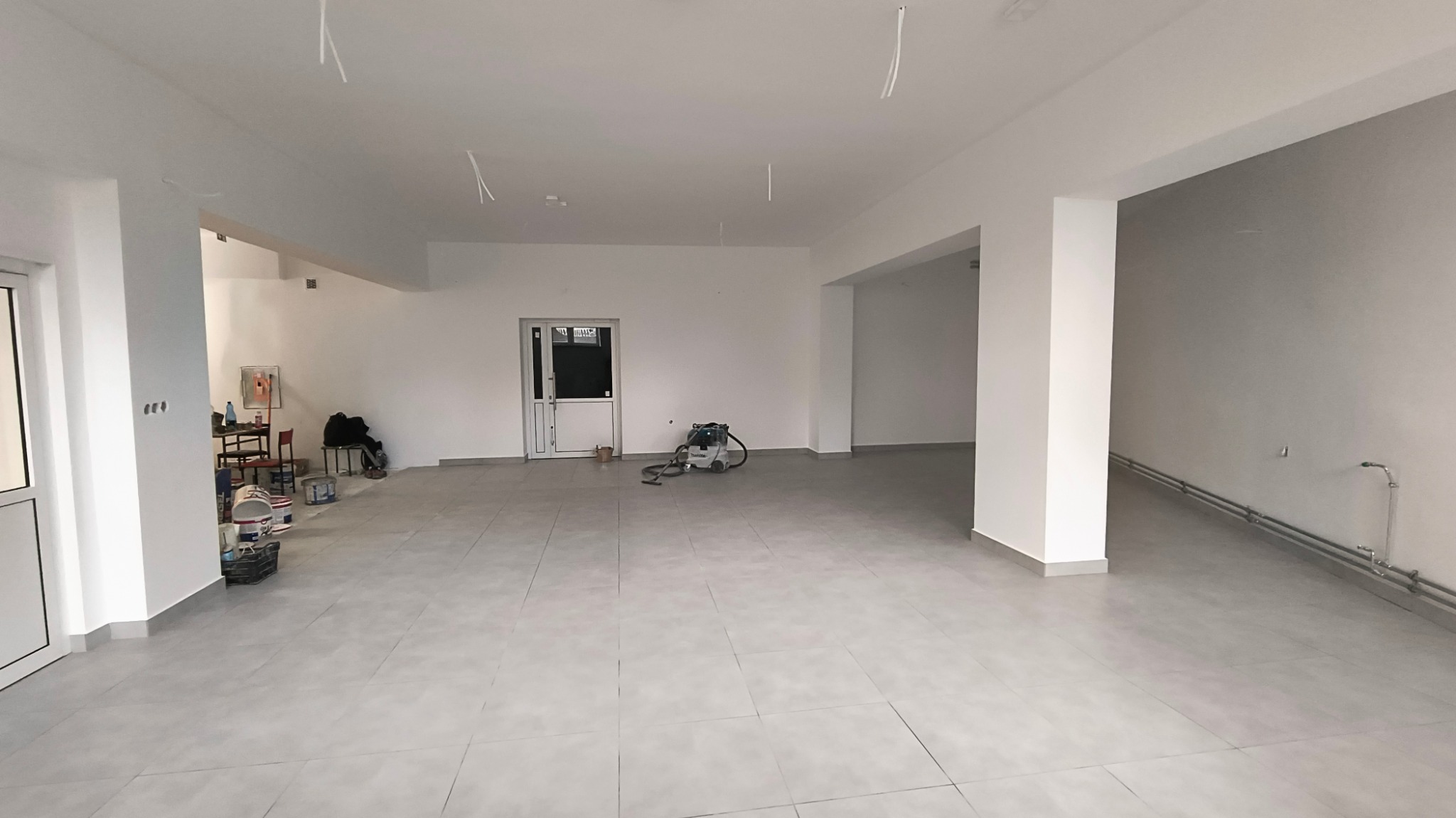 Przestronne, jasne pomieszczenie po remoncie z białymi ścianami i szarą podłogą z płytek. Widoczne ślady po pracach remontowych, w tym przewody zwisające z sufitu, odkurzacz przemysłowy...