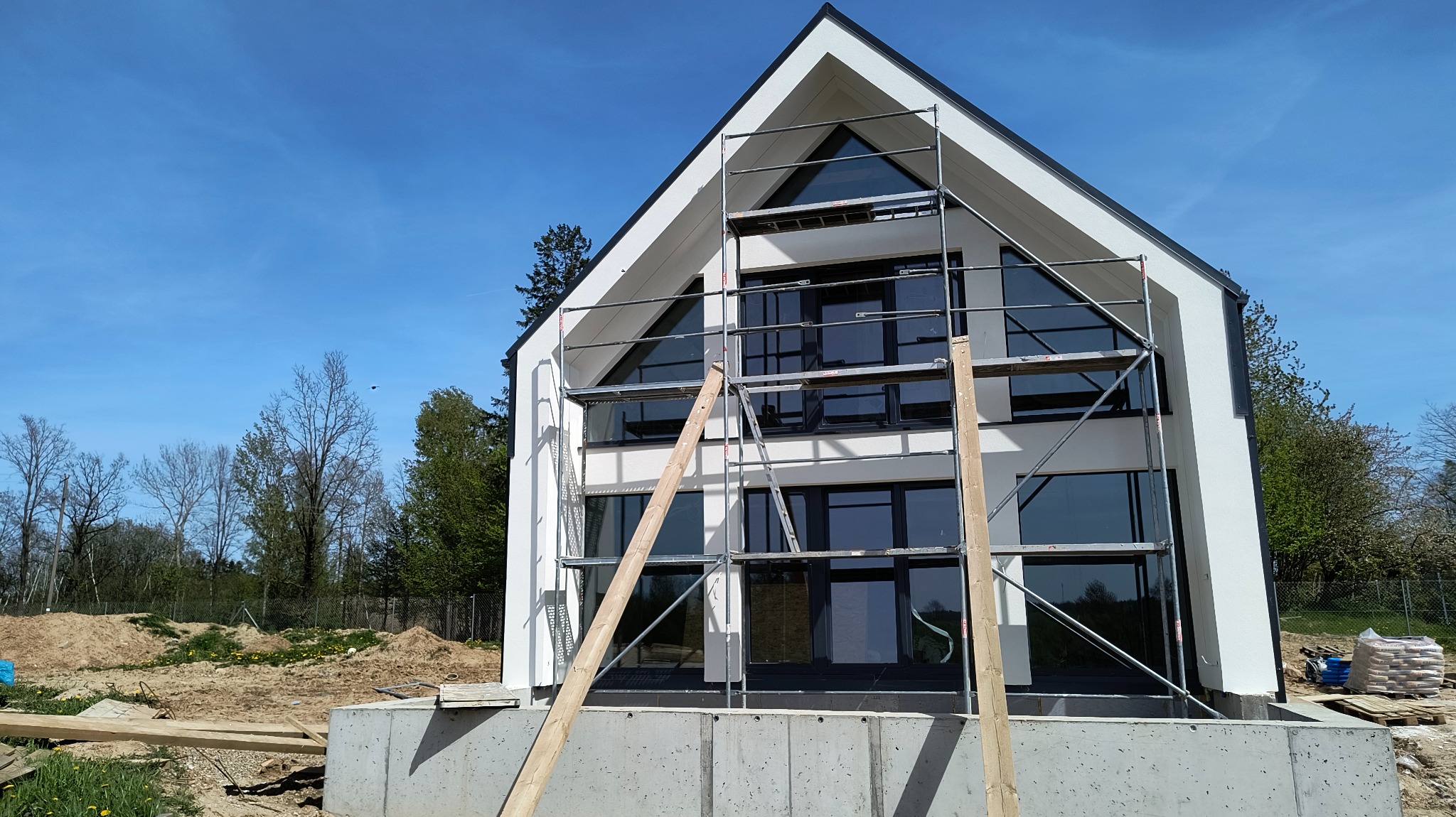 Nowoczesny dom jednorodzinny z dużymi oknami i rusztowaniem na elewacji, widok na budynek w trakcie prac wykończeniowych na zewnątrz, w tle błękitne niebo.