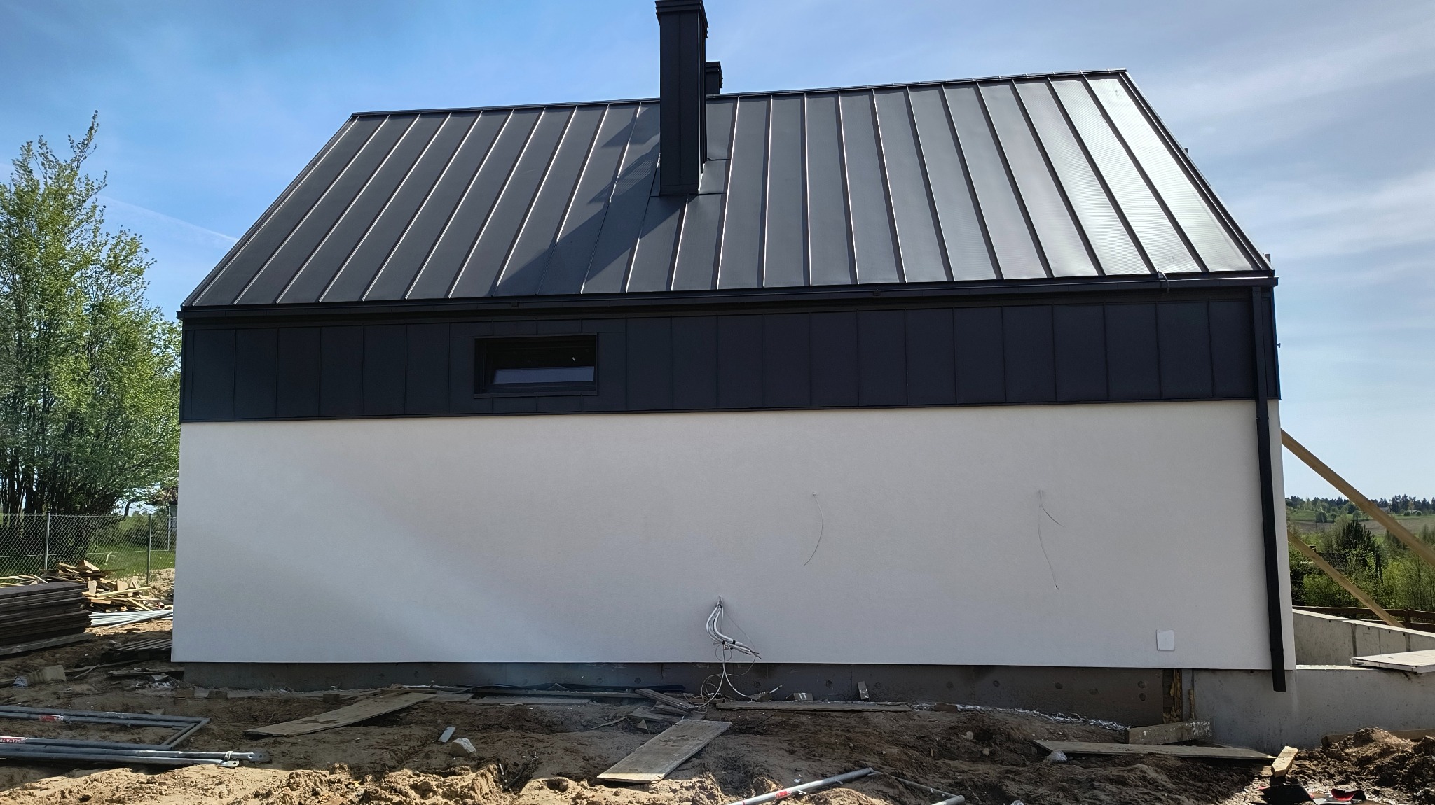 Elewacja domu jednorodzinnego z czarnym dachem z blachy na rąbek stojący i jasną, gładką ścianą, widoczne wystające kable i elementy instalacji w ziemi.