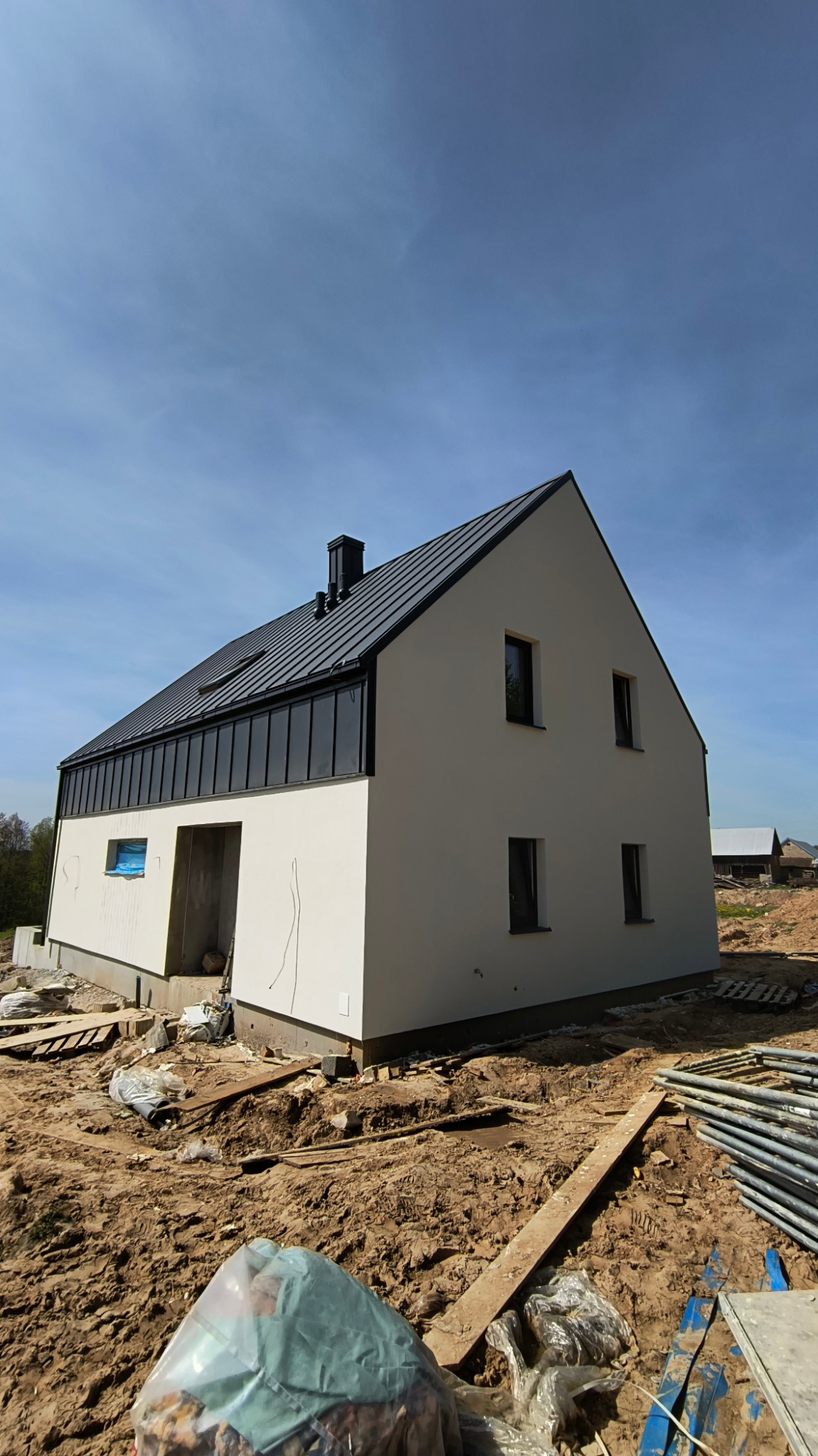 Surowy stan domu jednorodzinnego z ciemnym dachem i jasną elewacją, widoczny plac budowy z ziemią i materiałami.