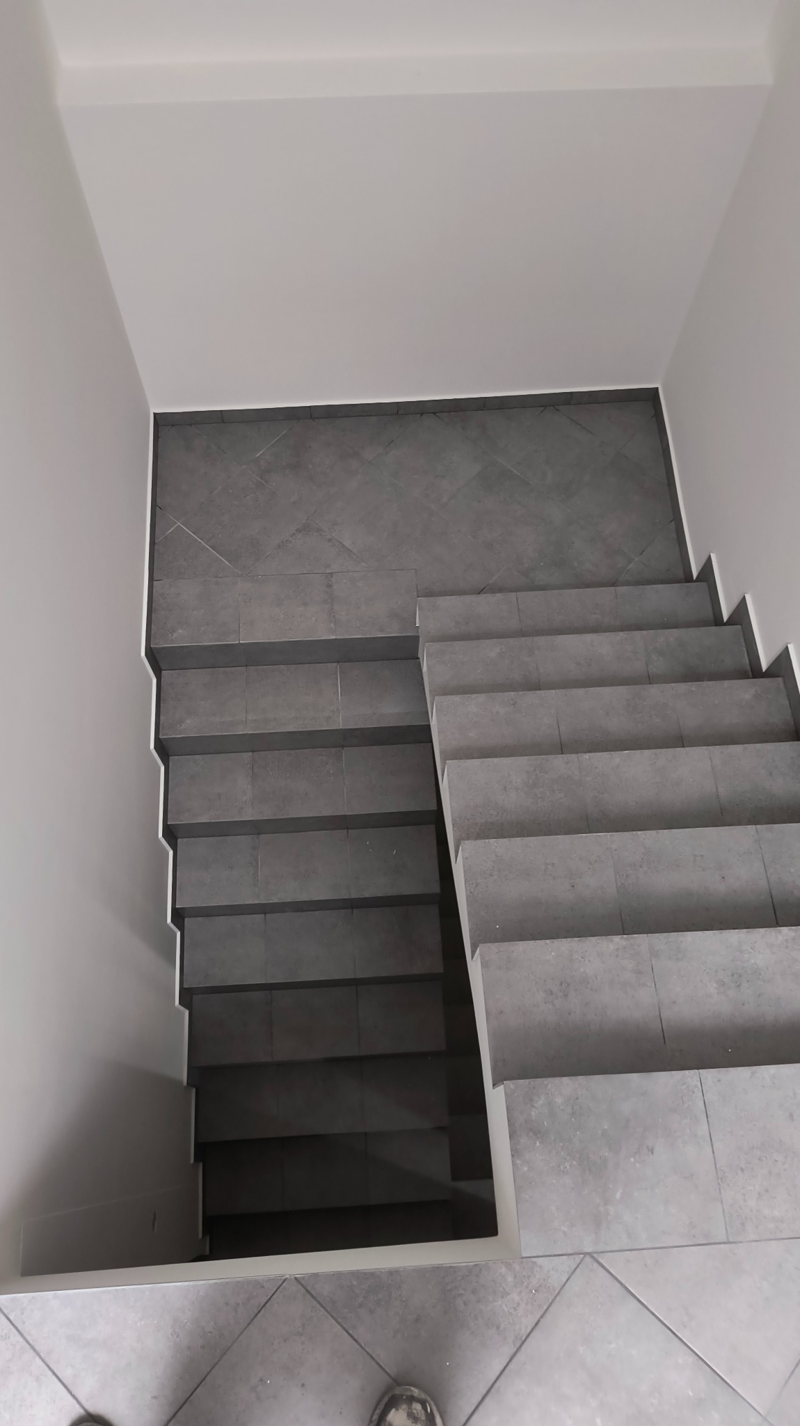 Schody wyłożone szarymi, kwadratowymi płytkami, widok z góry, białe ściany, minimalistyczny design.