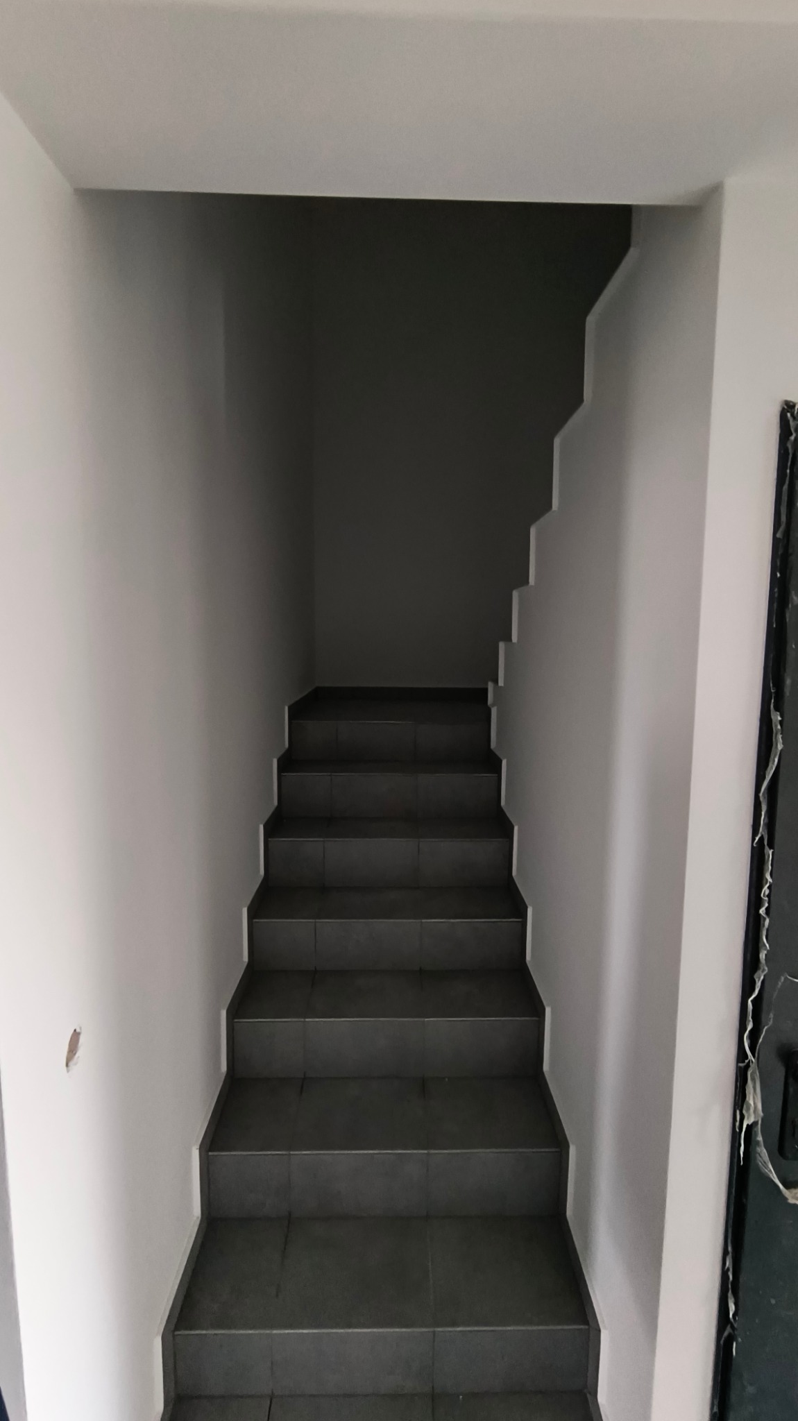 Schody wewnętrzne wyłożone ciemnymi płytkami, prowadzące w górę, ściany pomalowane na jasny odcień szarości, widoczny fragment uszkodzonej ściany i obudowy drzwi.