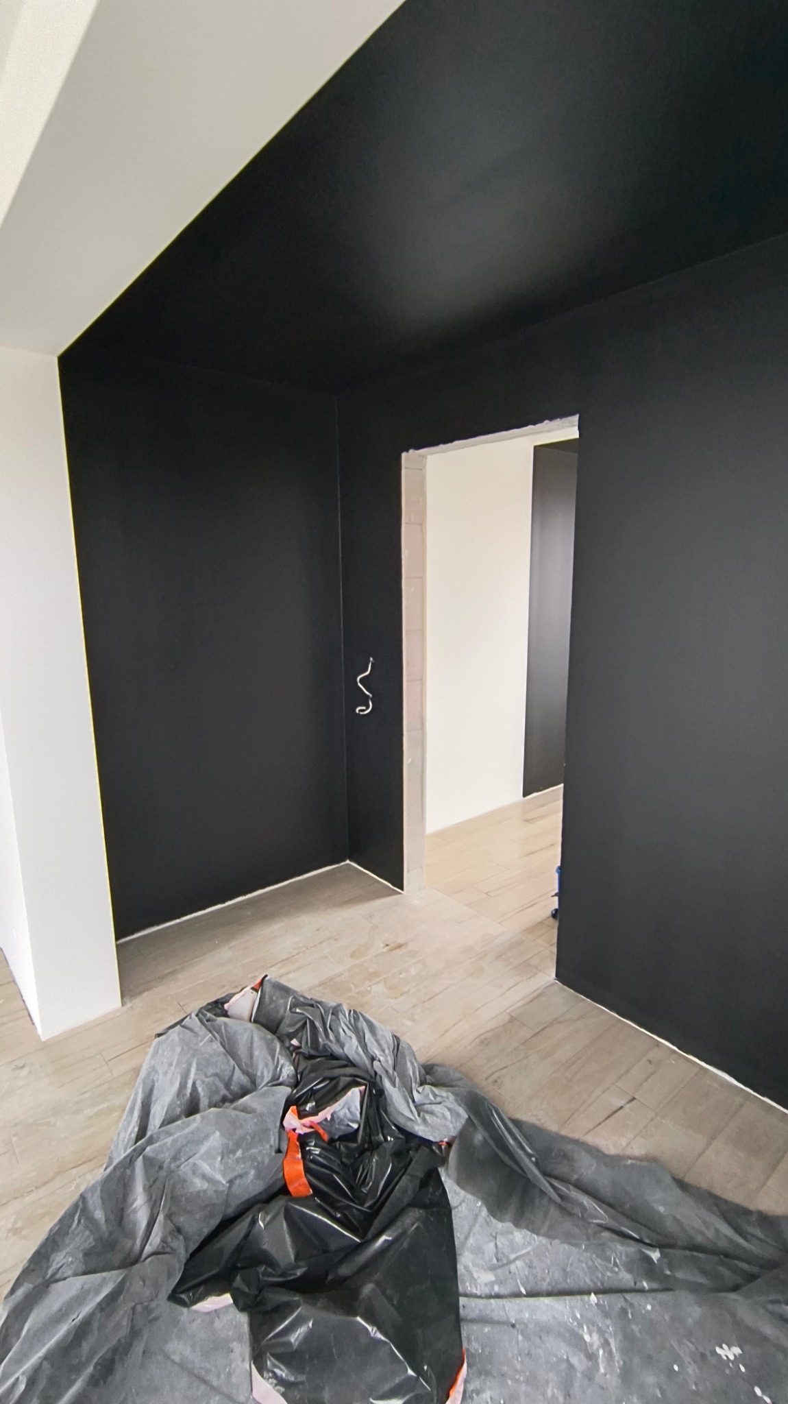 Pomieszczenie w trakcie malowania: czarny sufit i ściany, zabezpieczona podłoga folią malarską, widoczne przewody elektryczne wystające ze ściany.