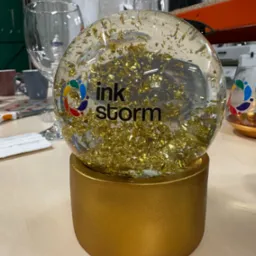 Szklana kula śnieżna z logo 'ink storm' i złotymi płatkami wewnątrz, umieszczona na złotym postumencie, na tle biurowym z widocznymi szklankami i innymi przedmiotami.