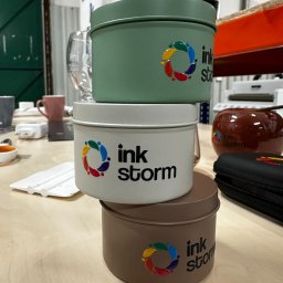 InkStorm - Personalizacja odzieży oraz gadżetów - Trzy metalowe puszki, jedna na drugiej, w odcieniach zieleni, bieli i brązu, z nadrukowanym logo 'ink storm' i kolorowym okręgiem z kropli, stojące na jasnym stole w warsztatowym otoczeniu.