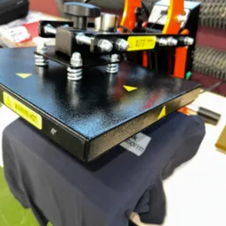 Proces nadruku na ciemnej koszulce za pomocą termoprasy, widoczna część koszulki pod płytą grzewczą i detale urządzenia, w tym ostrzeżenie o wysokiej temperaturze.