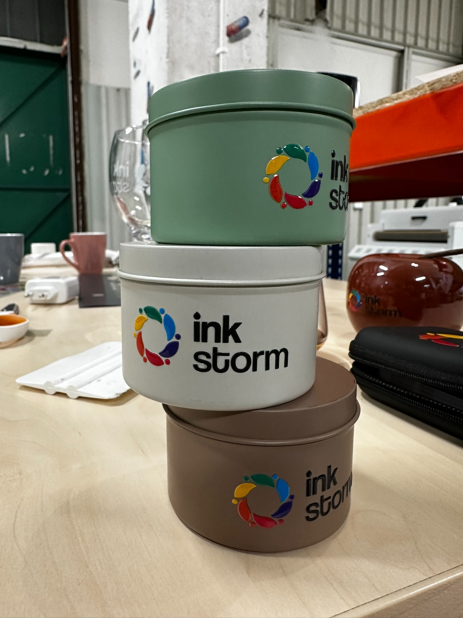 Trzy metalowe puszki, jedna na drugiej, w odcieniach zieleni, bieli i brązu, z nadrukowanym logo 'ink storm' i kolorowym okręgiem z kropli, stojące na jasnym stole w warsztatowym otoczeniu.