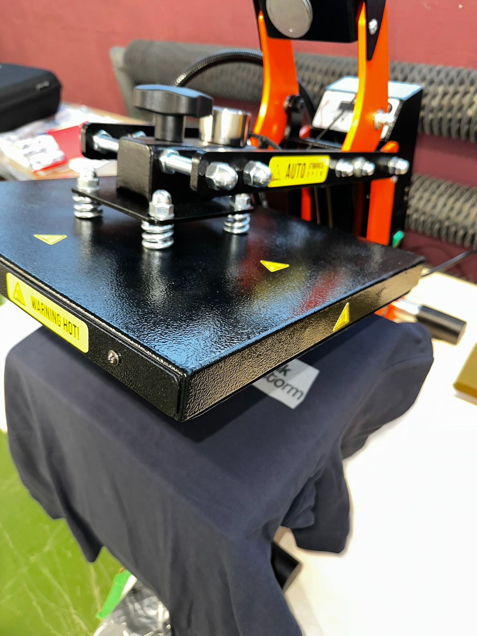 Proces nadruku na ciemnej koszulce za pomocą termoprasy, widoczna część koszulki pod płytą grzewczą i detale urządzenia, w tym ostrzeżenie o wysokiej temperaturze.