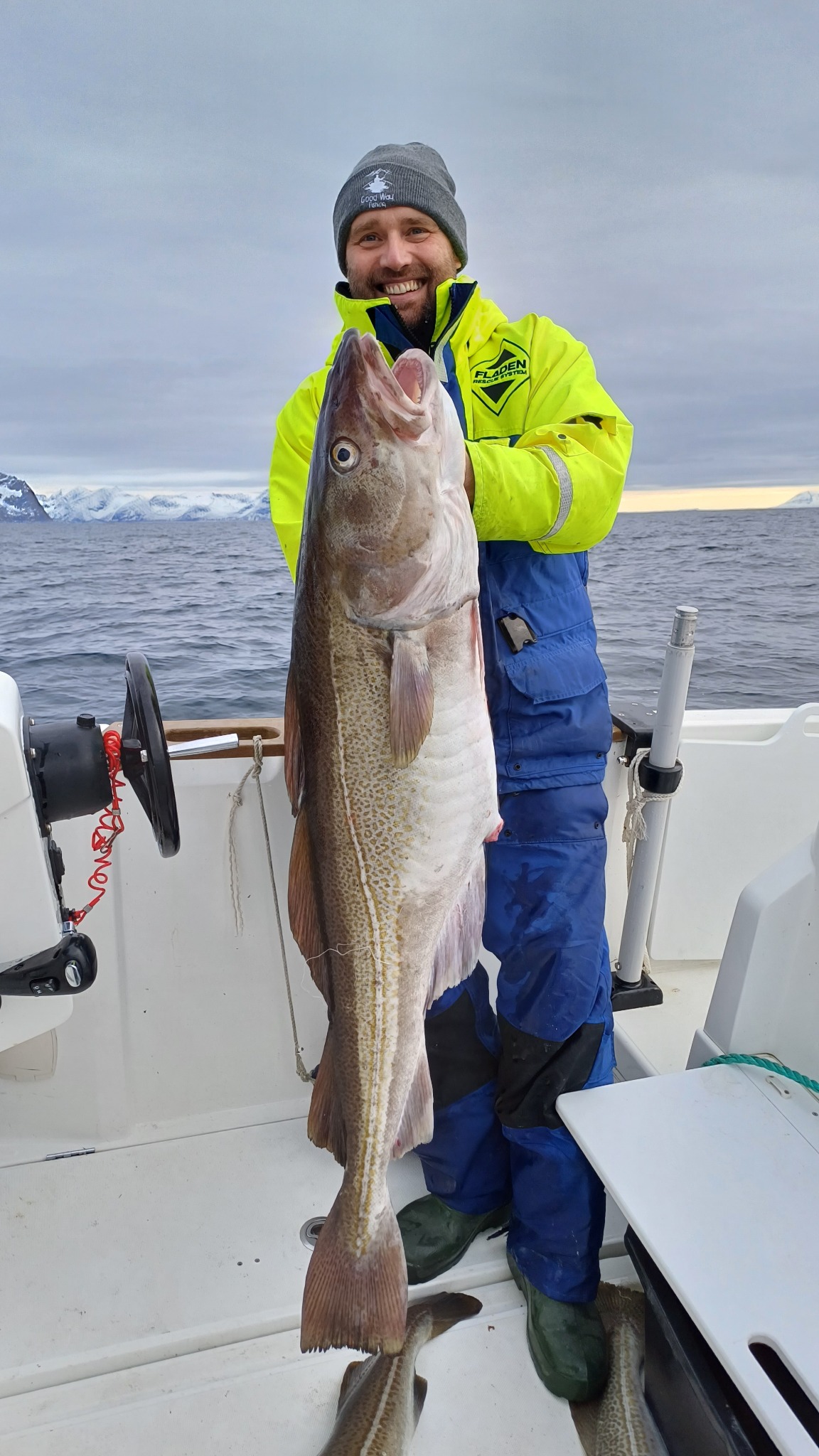Uśmiechnięty mężczyzna w neonowej kurtce i czapce trzyma dużego dorsza na pokładzie łodzi, w tle woda i ośnieżone góry.