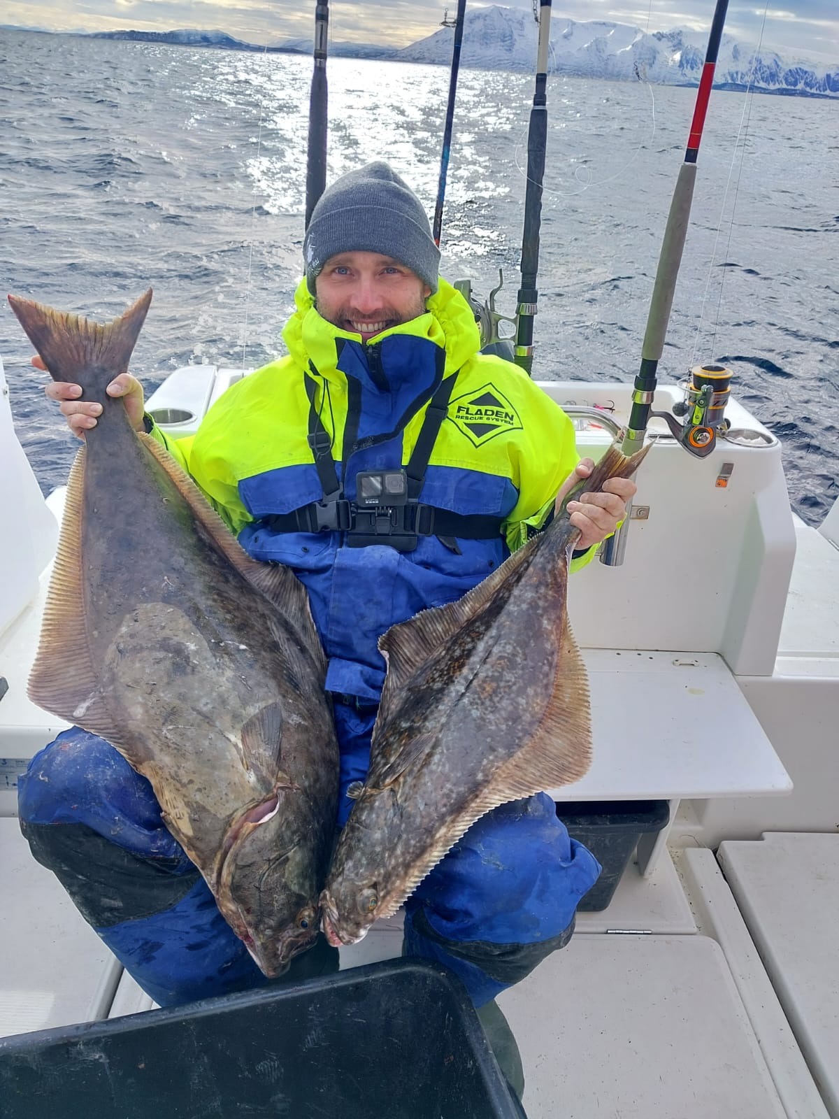 Uśmiechnięty mężczyzna w czapce i neonowej kurtce trzyma dwa duże halibuty na tle morza i górskich szczytów podczas wyprawy wędkarskiej.