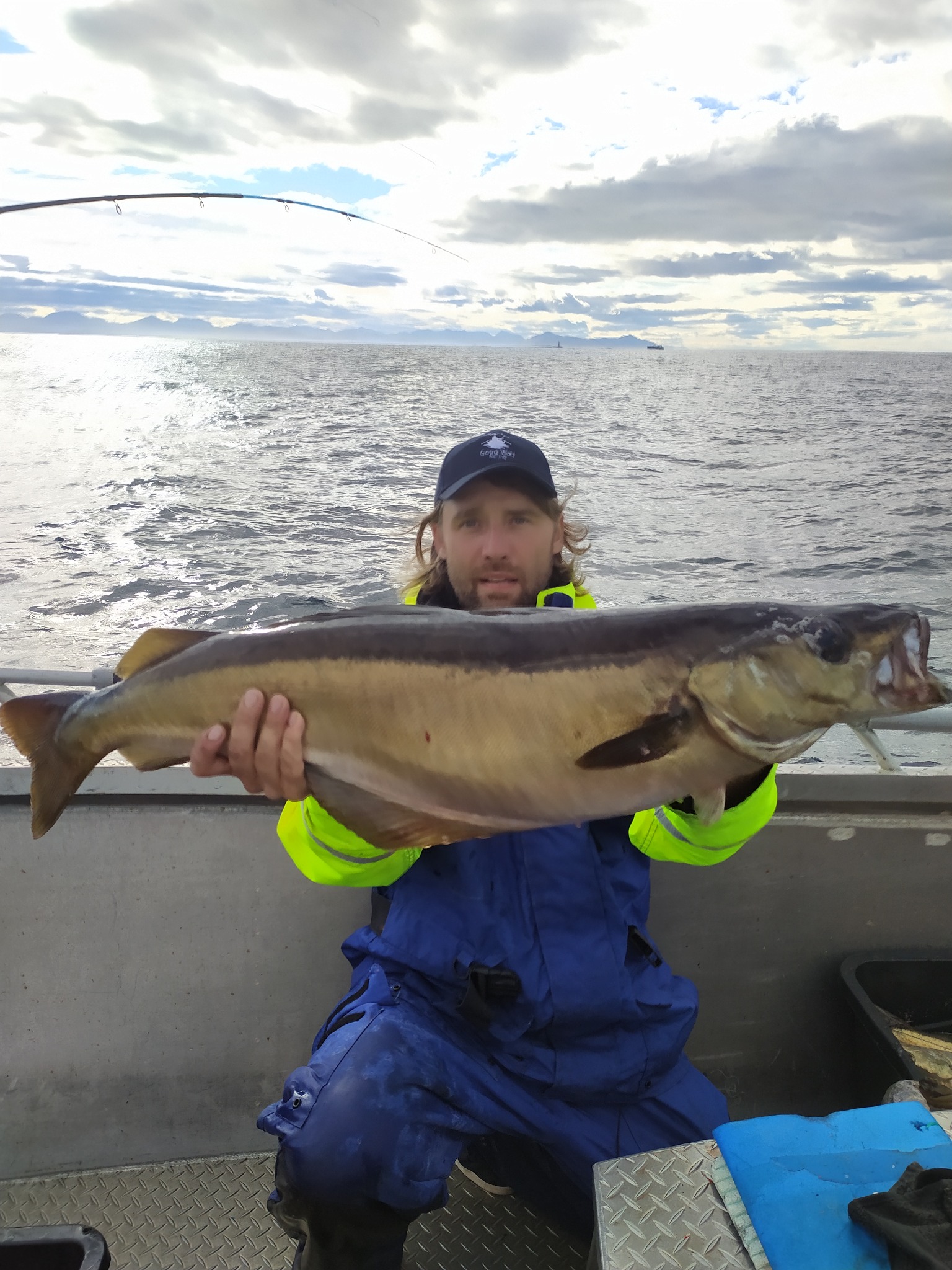 Wędkarz w niebieskim kombinezonie i czapce trzyma dużą rybę na tle morza i pochmurnego nieba, widoczna wędka.