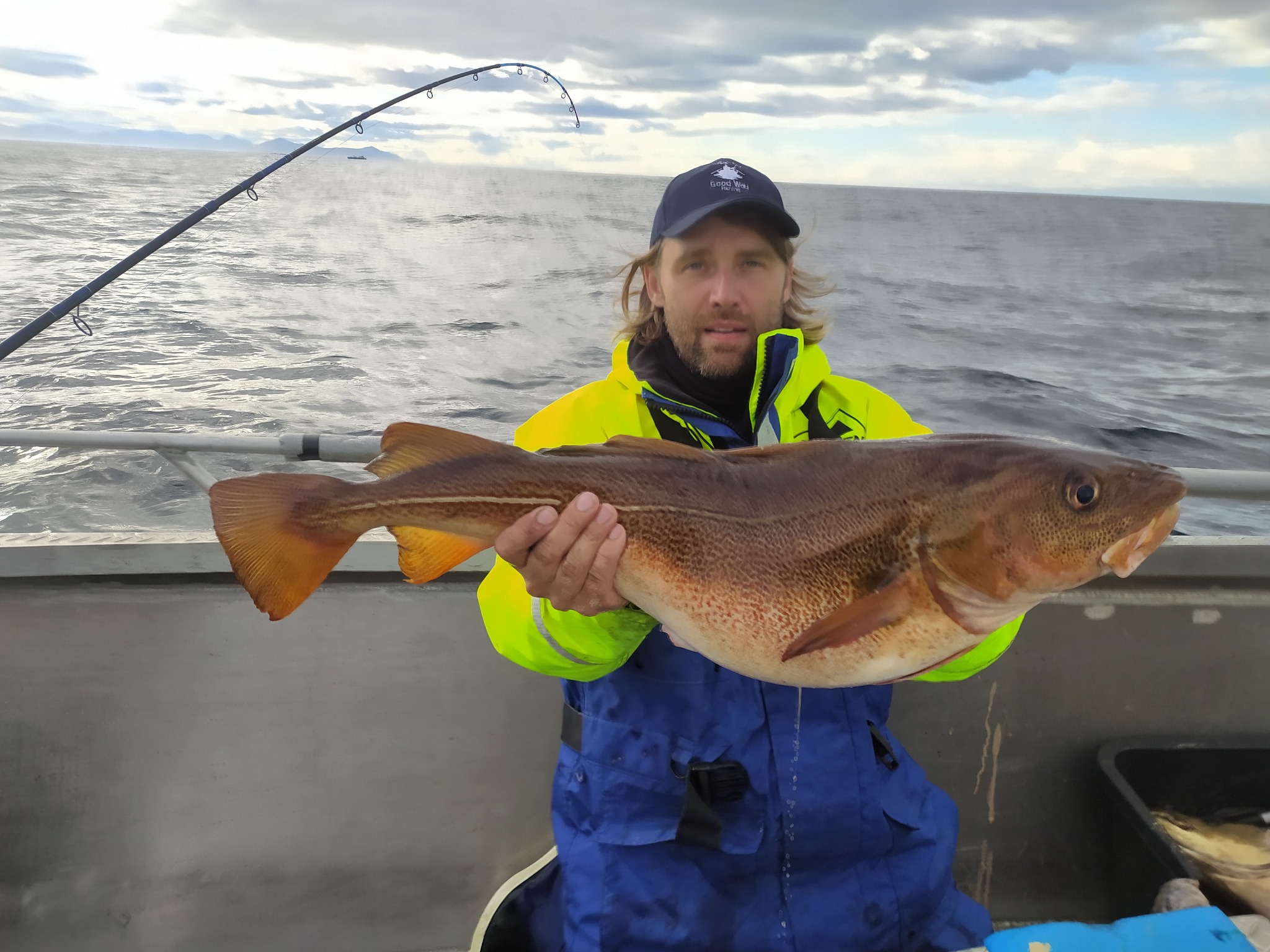 Wędkarz z dużą rybą (dorsz) na tle morza i pochmurnego nieba, w tle widoczna wędka.