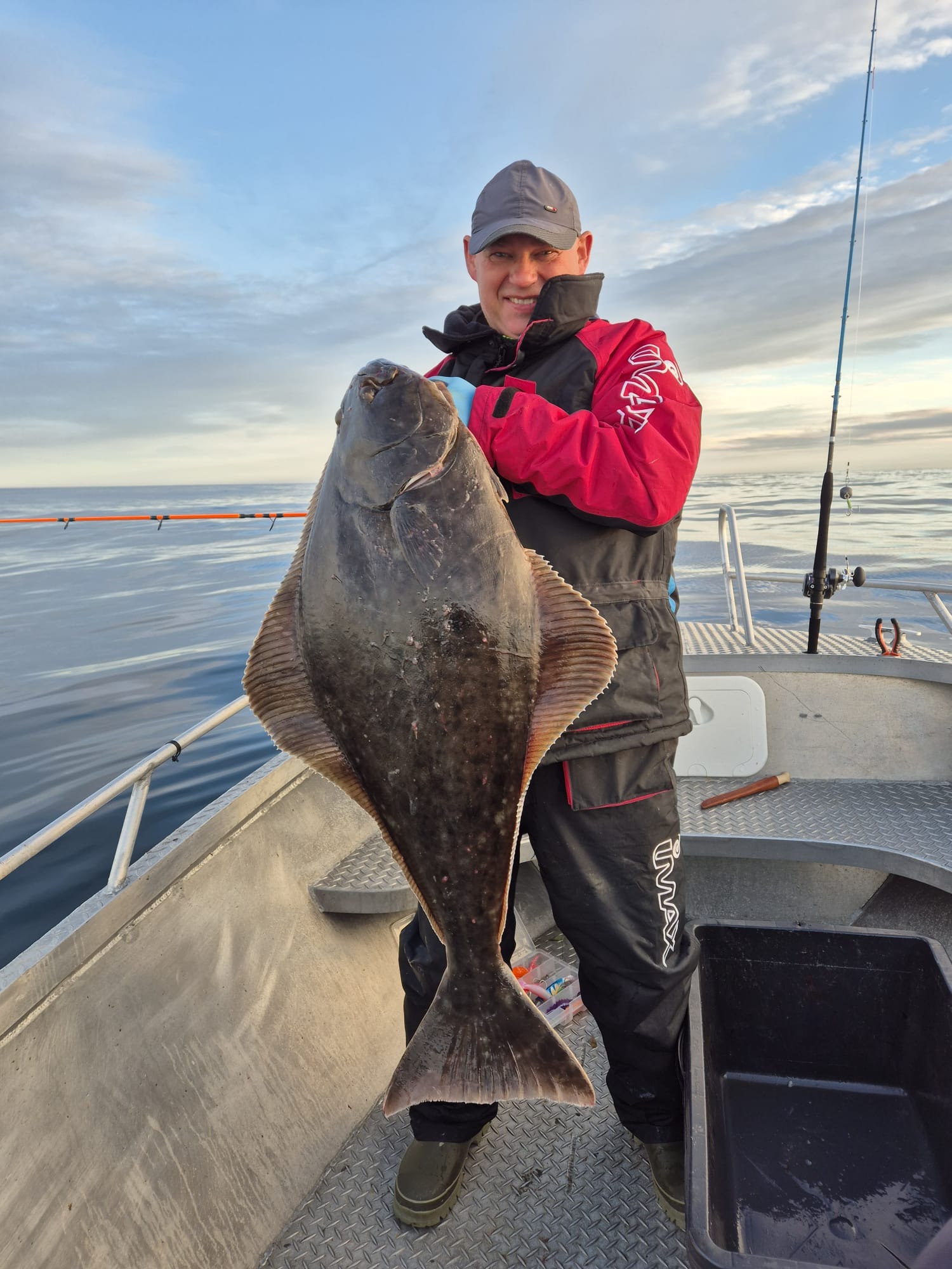 Uśmiechnięty mężczyzna w kurtce rybackiej prezentuje dużego halibuta na tle morza i nieba z chmurami, stojąc na łodzi.
