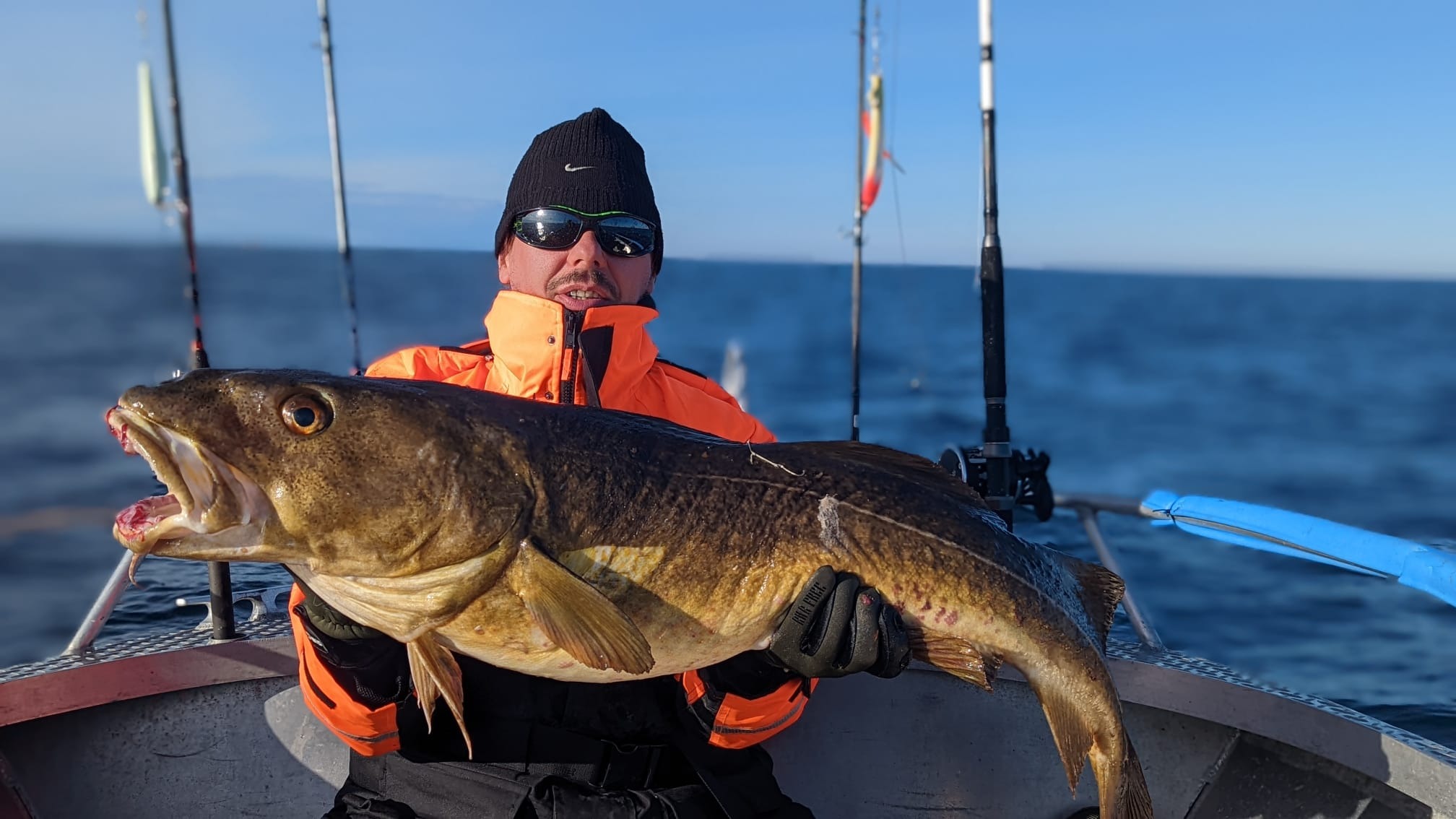 Mężczyzna w pomarańczowej kurtce i czarnej czapce trzyma ogromnego dorsza na tle morza i wędek.