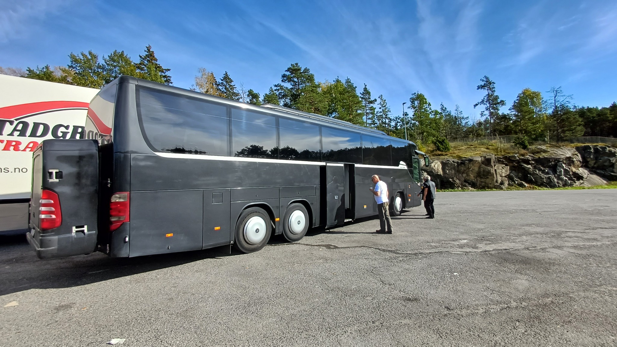 Czarny autokar turystyczny z otwartymi drzwiami, stojący na parkingu z dwoma osobami w pobliżu, na tle drzew i skał.