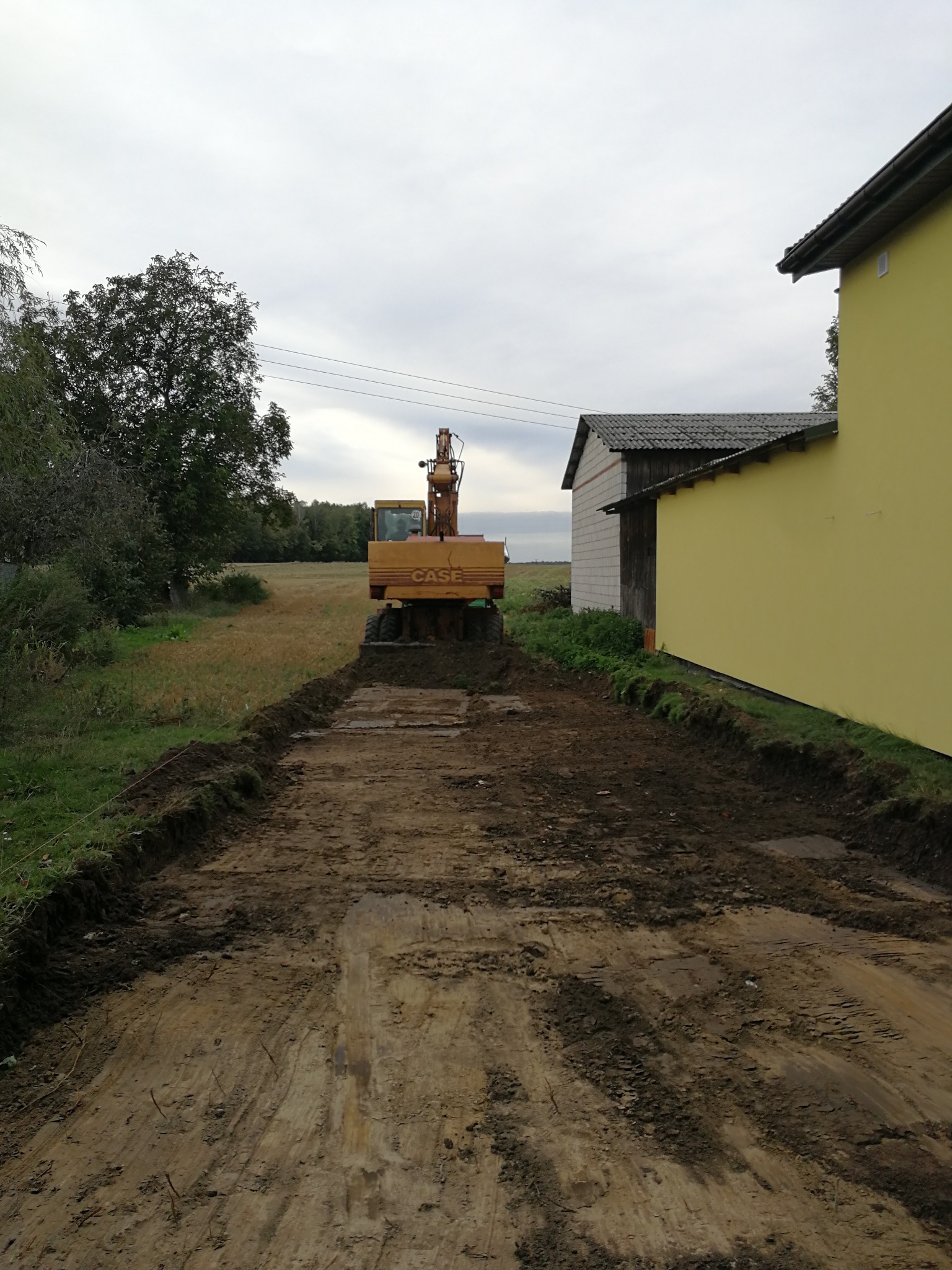 Żółta koparka CASE na wytyczonym, błotnistym terenie budowy, z widocznymi śladami opon i przygotowanymi krawędziami. W tle budynki i drzewa.