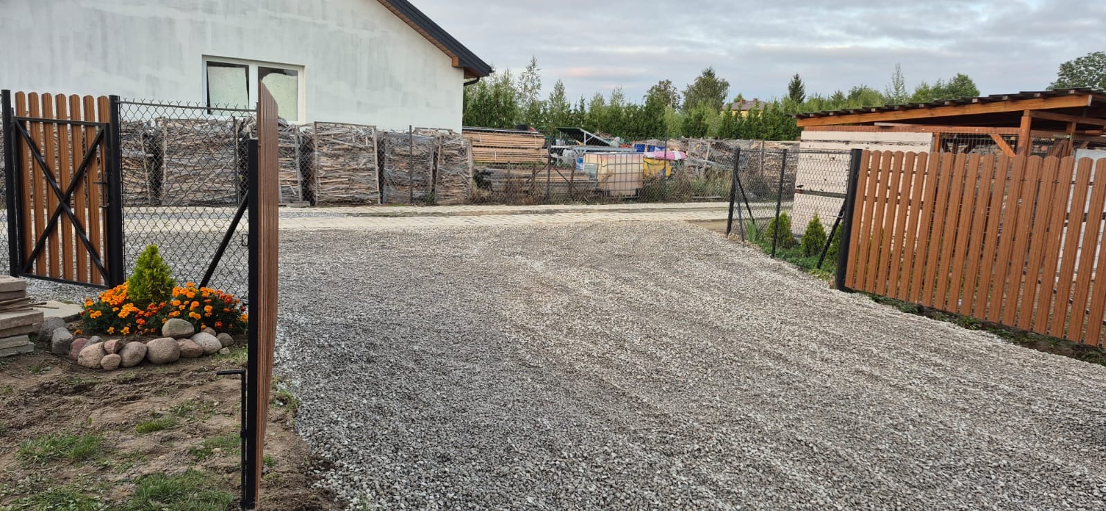 Utwardzona żwirem droga wjazdowa z drewnianą bramą i ogrodzeniem, obok domu z widocznymi materiałami budowlanymi za siatką. Na pierwszym planie rabata kwiatowa.