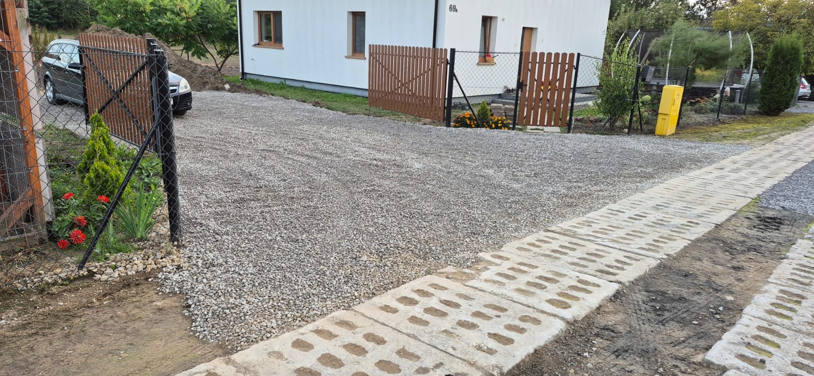 Utwardzony żwirem podjazd do domu z brązowym ogrodzeniem i betonowymi płytami ażurowymi po prawej stronie. Widoczny samochód i zieleń.