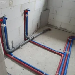 Instalacja wodna z rur PEX w kolorze niebieskim i czerwonym, zamocowana do ściany z betonu kom&oacute;rkowego. Szare rury kanalizacyjne łączą piony.