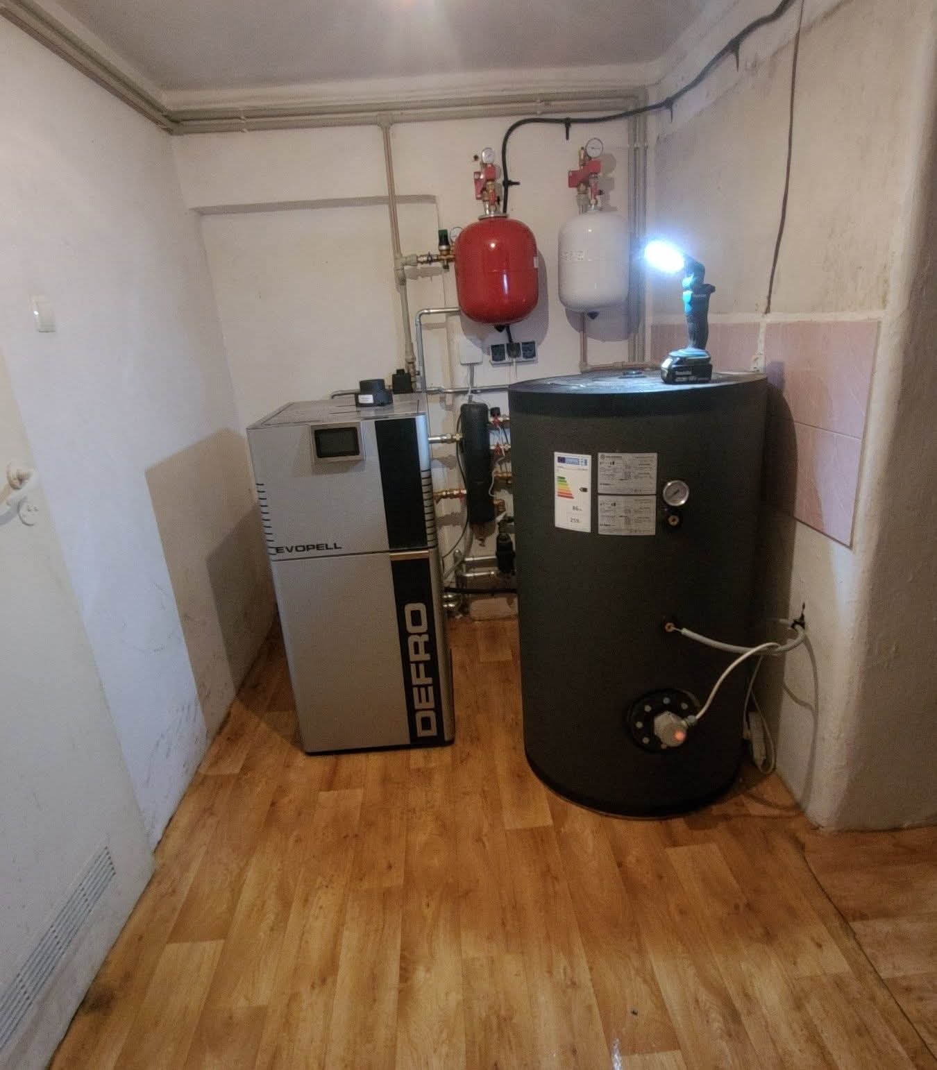 Zestaw grzewczy Defro z zasobnikiem i osprzętem w kotłowni. Na zasobniku akumulatorowa lampa warsztatowa. Widoczne rury, zawory i zbiorniki wyrównawcze.