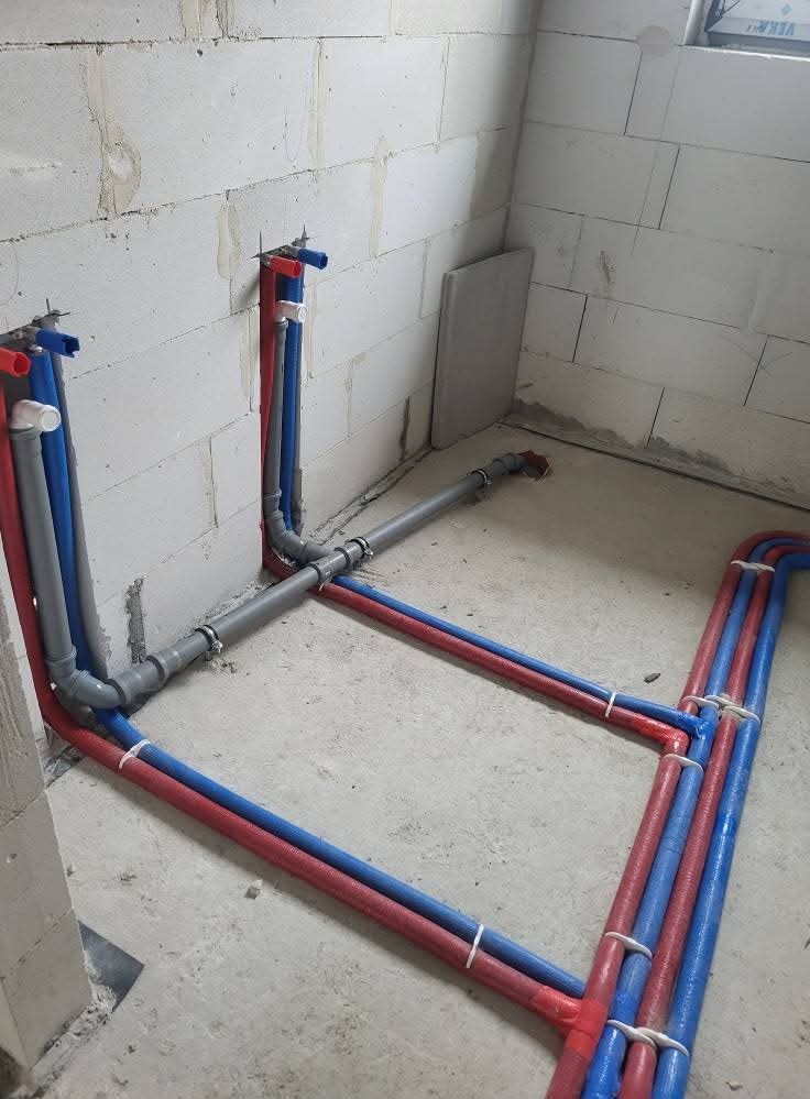 Instalacja wodna z rur PEX w kolorze niebieskim i czerwonym, zamocowana do ściany z betonu komórkowego. Szare rury kanalizacyjne łączą piony.
