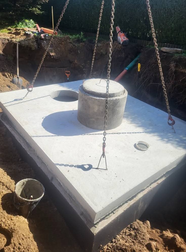 Betonowy szambo z pokrywą, opuszczany do wykopu za pomocą łańcuchów. Widoczne narzędzia i elementy instalacji w tle. Ujęcie z góry, w słoneczny dzień.