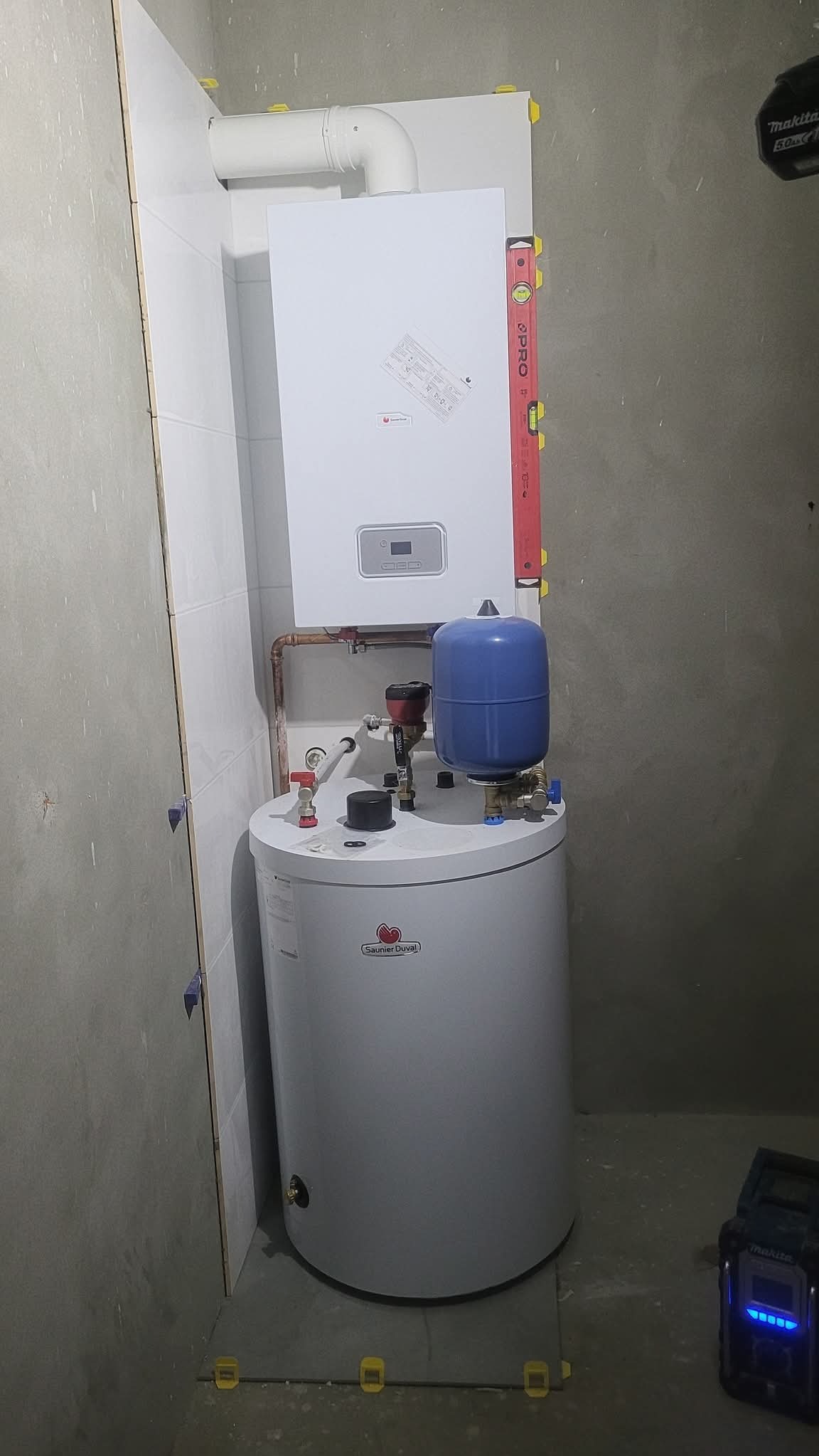 Biały piec CO i zasobnik ciepłej wody użytkowej zamontowane w kotłowni w Szemud. Poziomica przy piecu. Widoczne płytki i narzędzia.