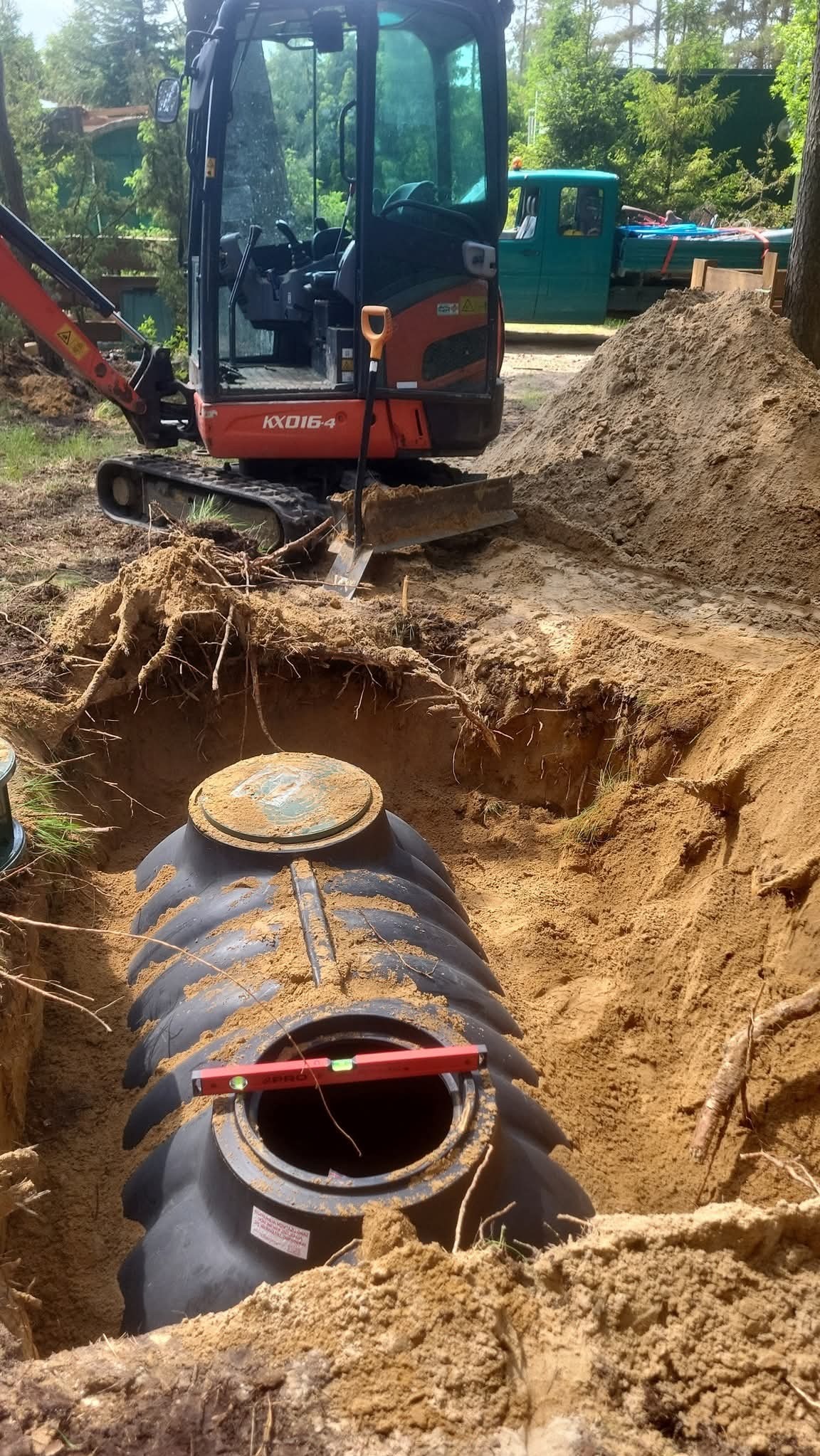 Montaż szamba betonowego w wykopie, z użyciem poziomicy i minikoparki Kubota KX016-4. Widoczny zielony samochód dostawczy w tle, teren zalesiony.