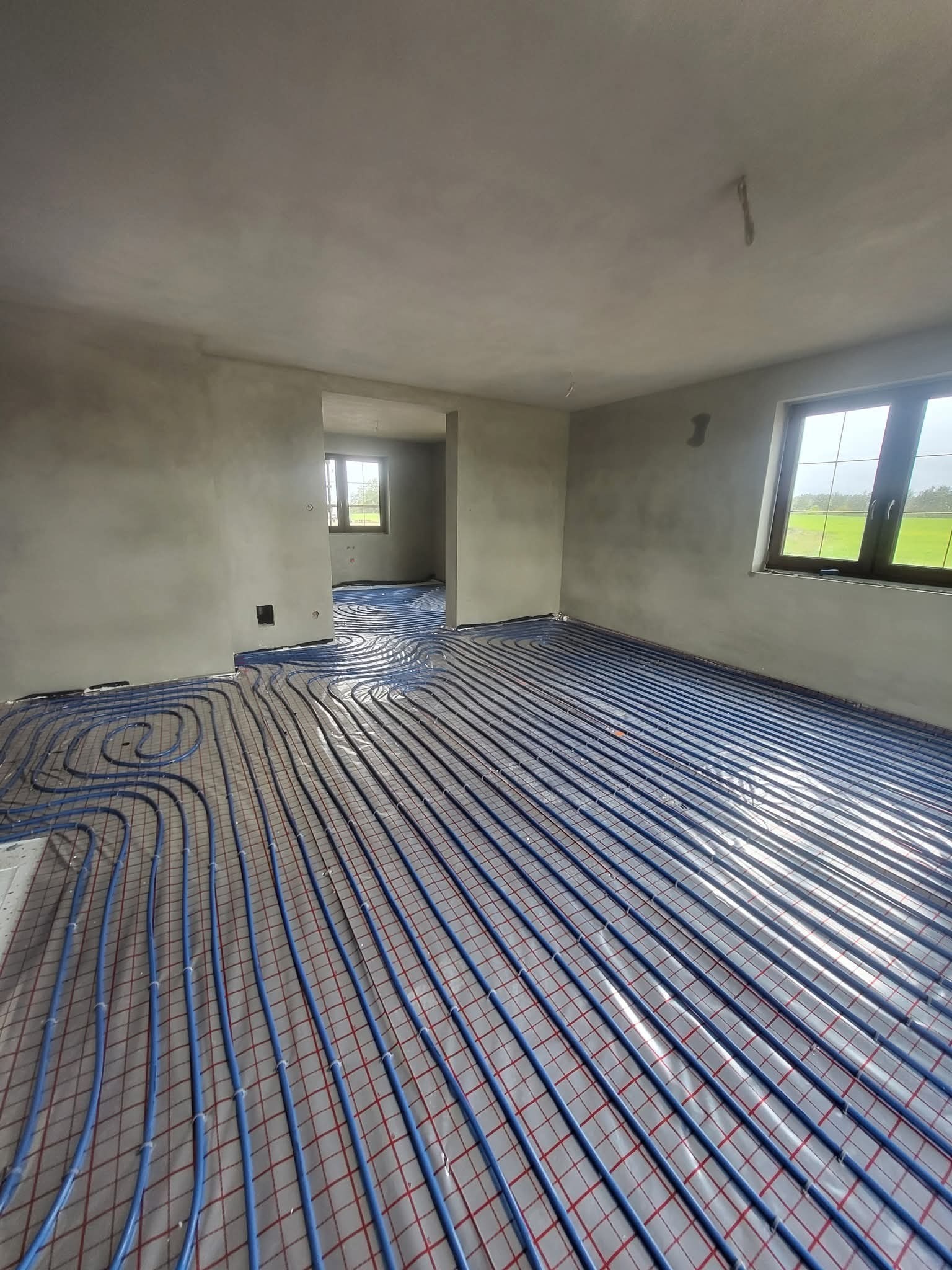 Niebieskie rury ogrzewania podłogowego ułożone na folii w surowym wnętrzu z oknem. Widok na sąsiednie pomieszczenie z identyczną instalacją.