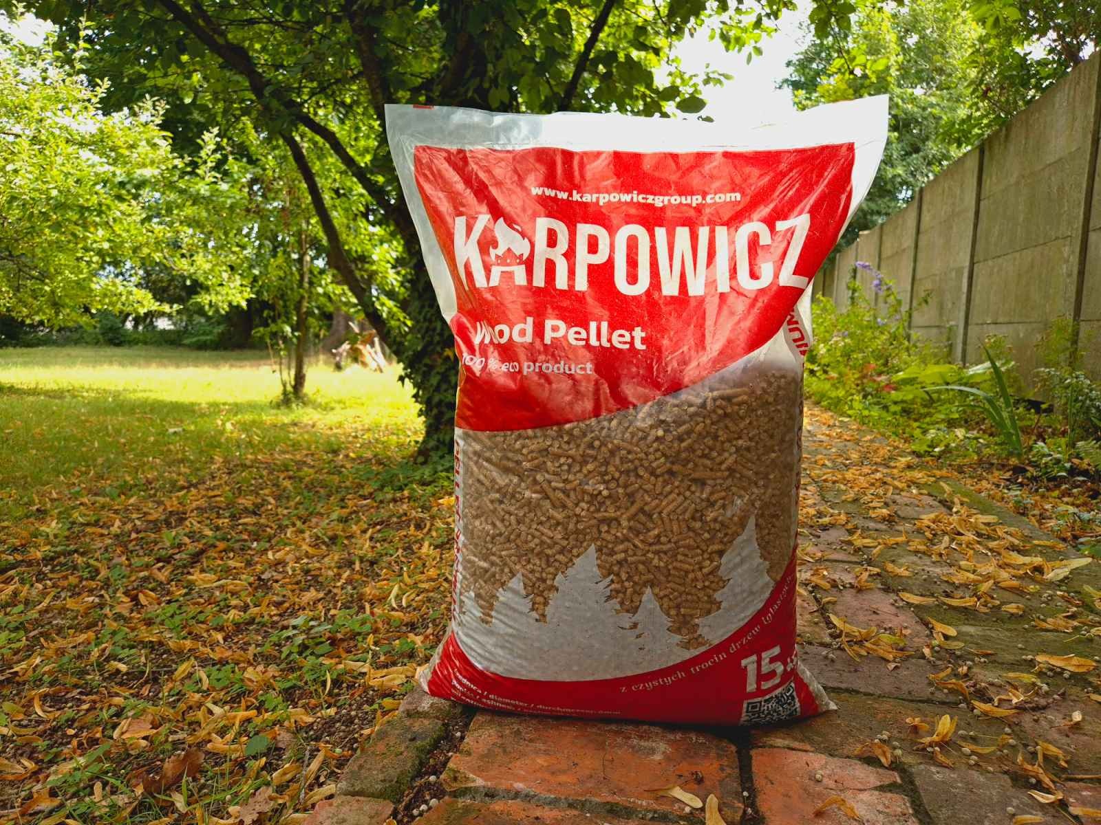 Worek z pelletem drzewnym marki Karpowicz, stojący na ceglanym murku w ogrodzie, z widocznymi liśćmi na trawie i betonowym ogrodzeniem w tle.