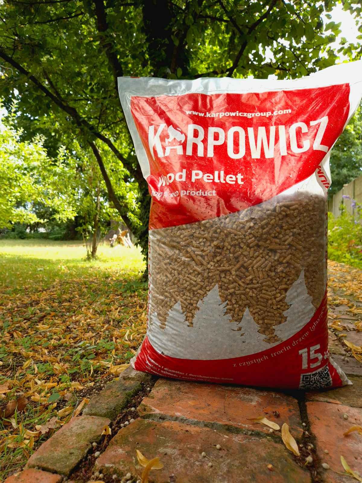 Worek wypełniony peletem drzewnym na tle ogrodu z drzewami i opadłymi liśćmi, widoczne logo producenta na czerwonym tle.