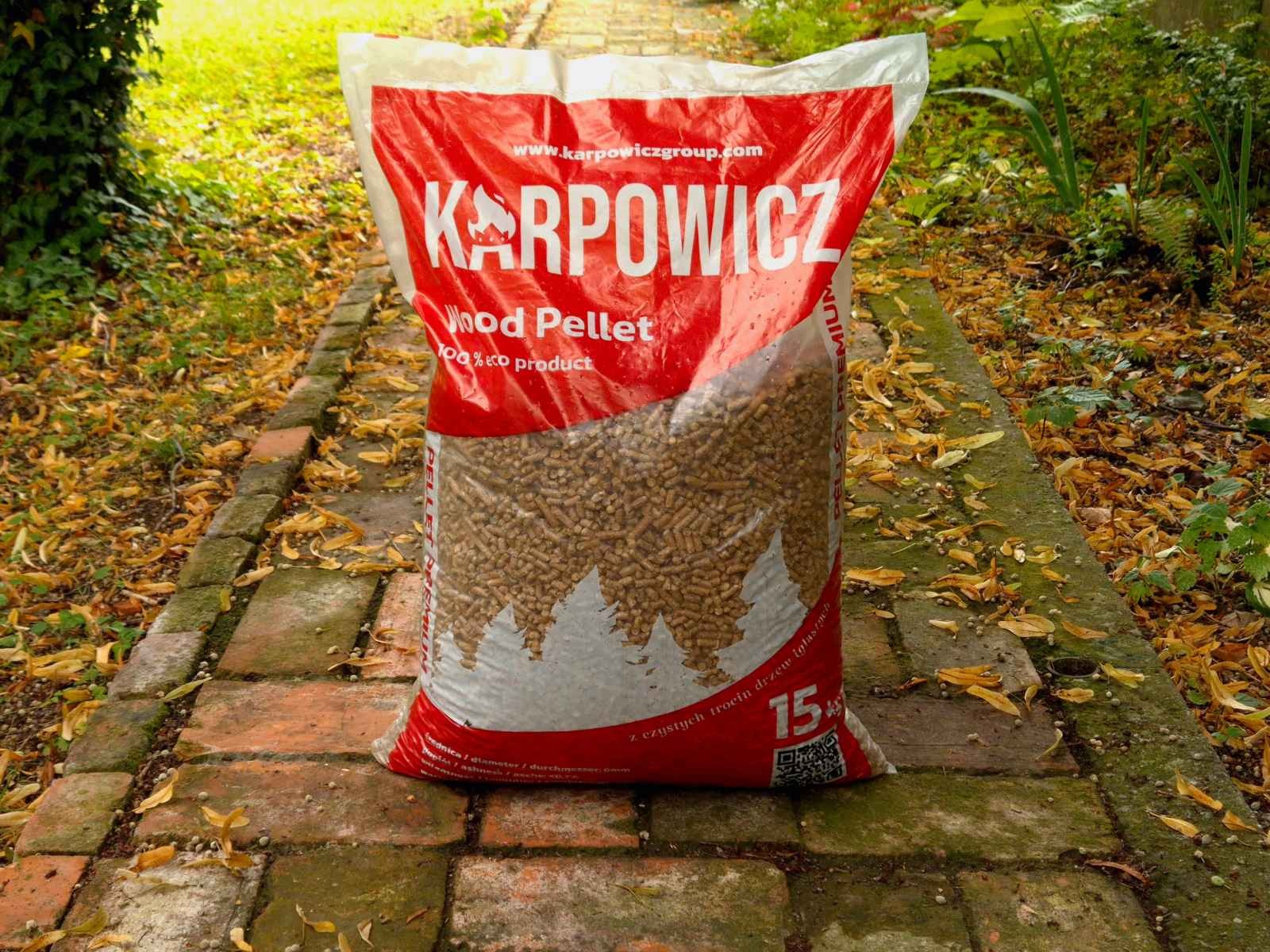 Worek pelletu drzewnego marki Karpowicz, ustawiony na ścieżce z cegieł, otoczony opadłymi liśćmi, z widocznym napisem '100% eco product' i wagą 15kg.