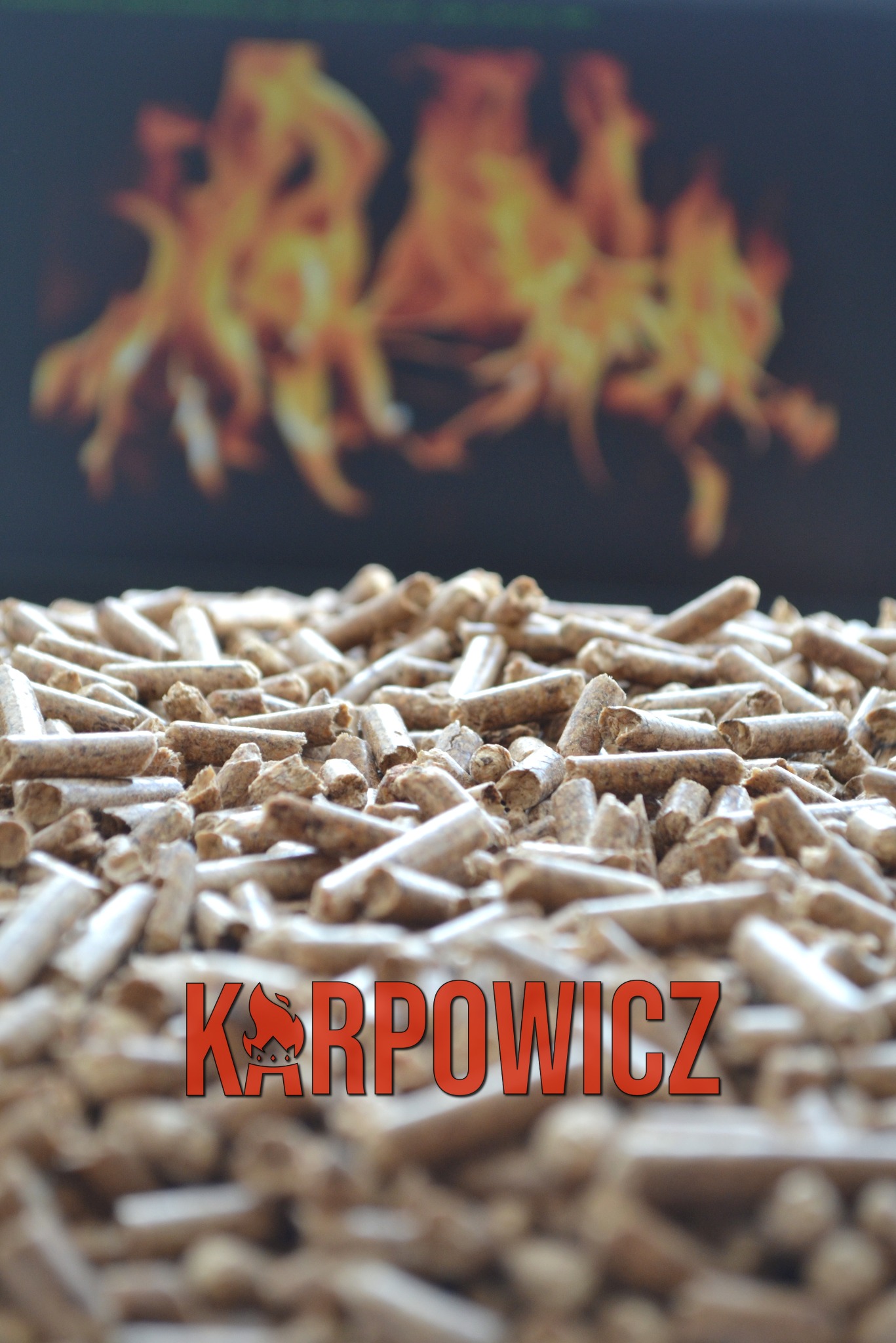 Stos pelletu drzewnego z widocznym logo firmy 'KARPOWICZ' na pierwszym planie, w tle rozmyte płomienie ognia.