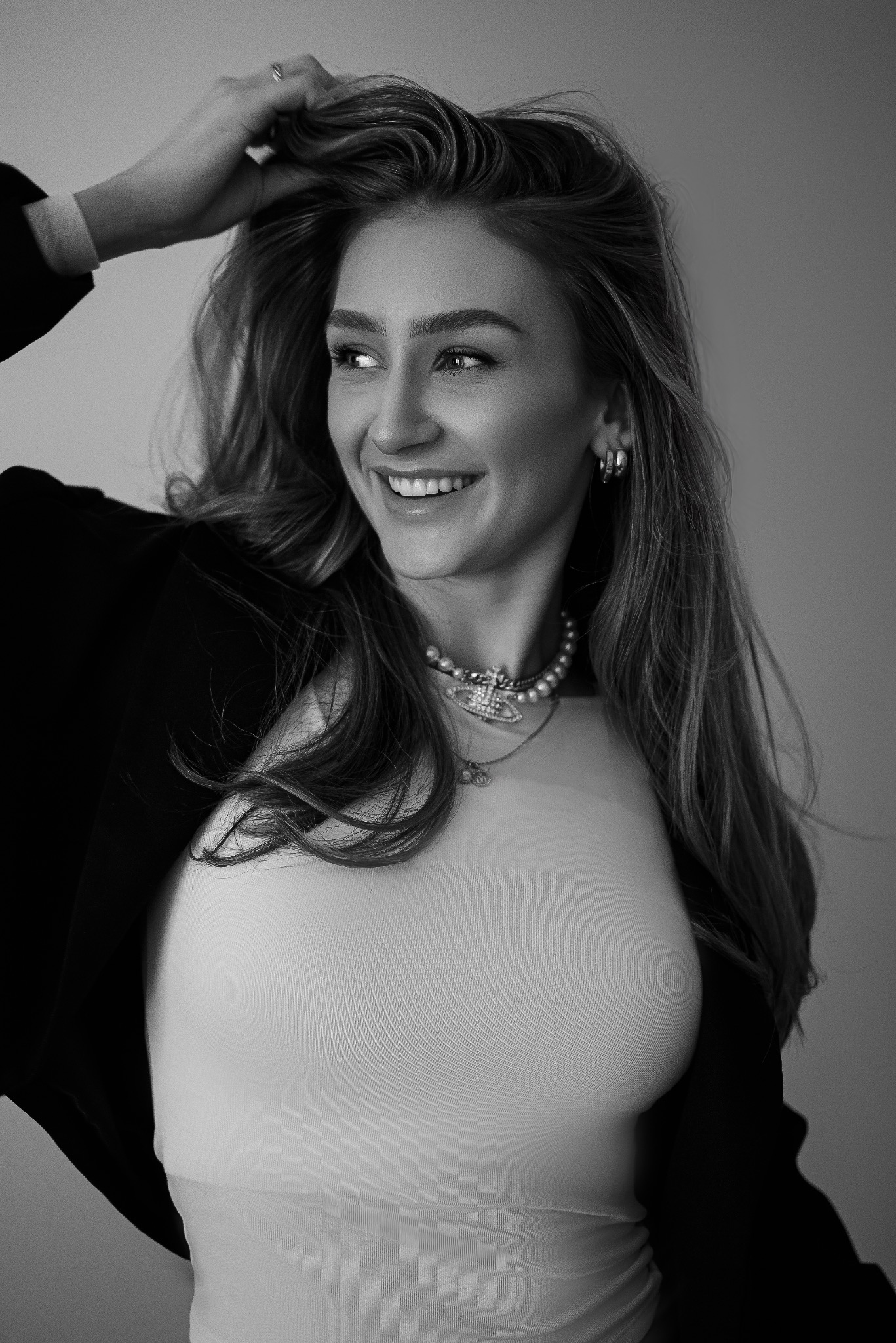 Portret uśmiechniętej kobiety z długimi włosami, w czarnej marynarce i z biżuterią na szyi, odwróconej w bok, z ręką we włosach, na jasnym tle.