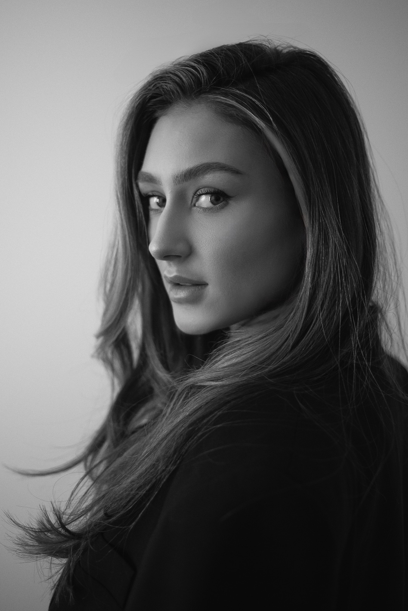Czarno-biały portret kobiety z długimi, pofalowanymi włosami, odwróconej przez ramię, z delikatnym makijażem i poważnym spojrzeniem.
