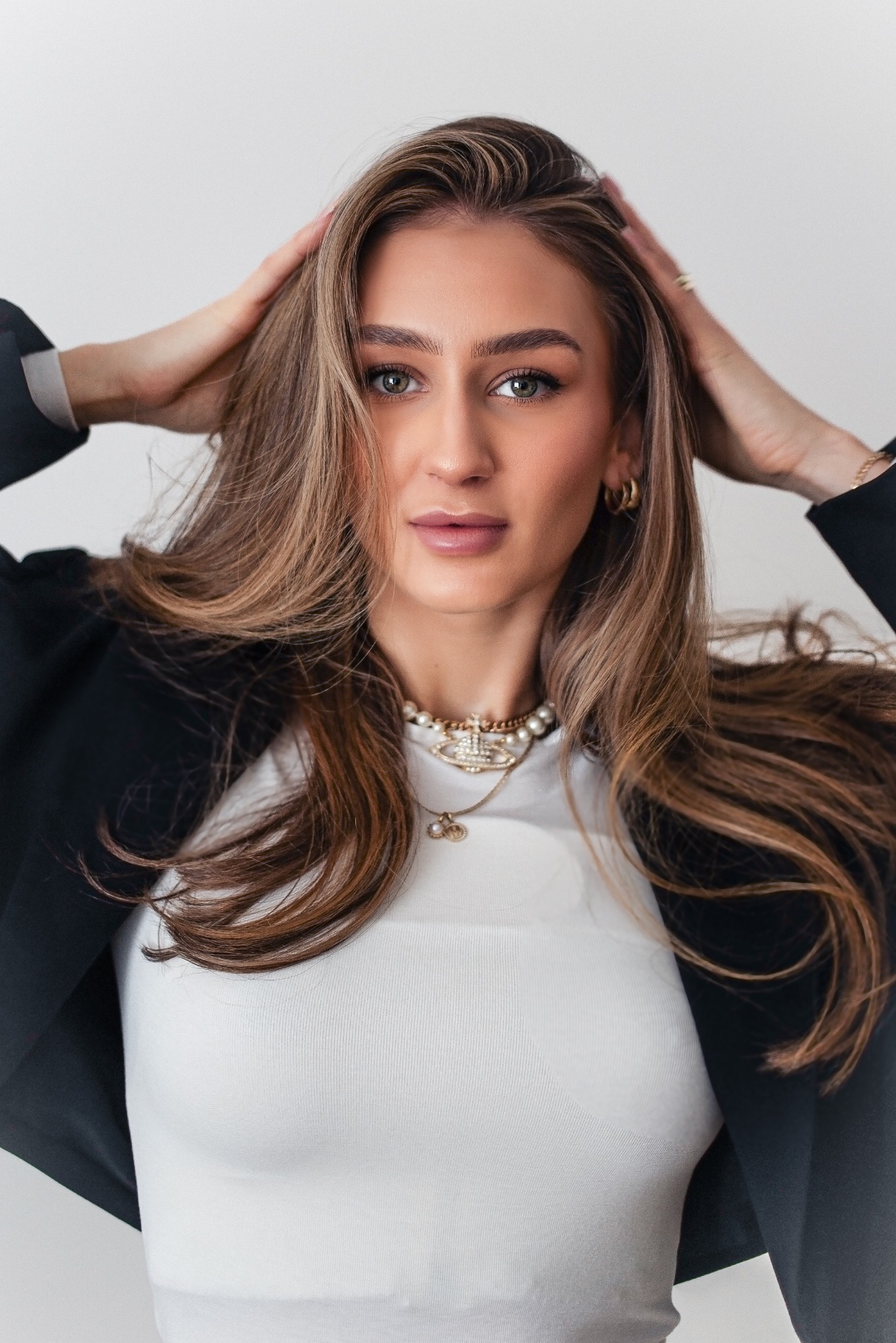 Portret kobiety z długimi brązowymi włosami, ubranej w białą bluzkę i czarną marynarkę, pozującej z rękami uniesionymi do góry, z delikatnym makijażem i biżuterią.