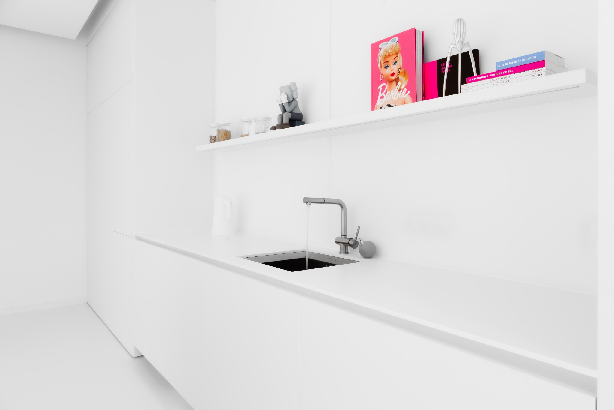 Minimalistyczna, całkowicie biała kuchnia z płynącą wodą z kranu, na półce dekoracje: słoiki, figurka, książka o Barbie i inne książki.